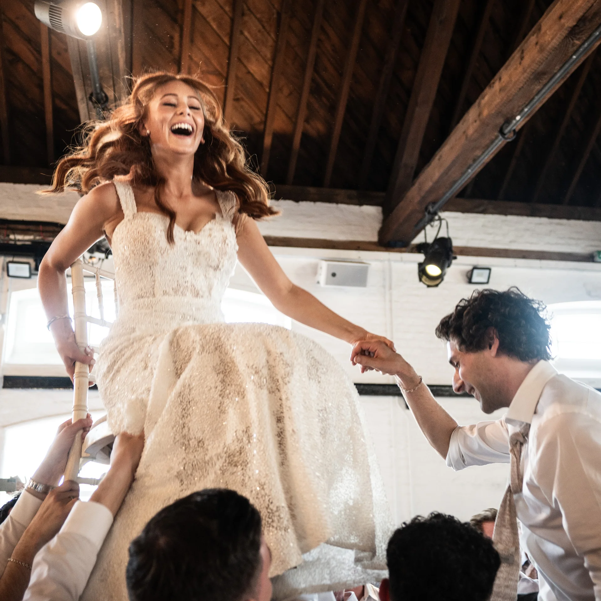 A joyful bride laughs aloud as she is lifted high on a chair during the wedding reception celebration under exposed wooden rafters.