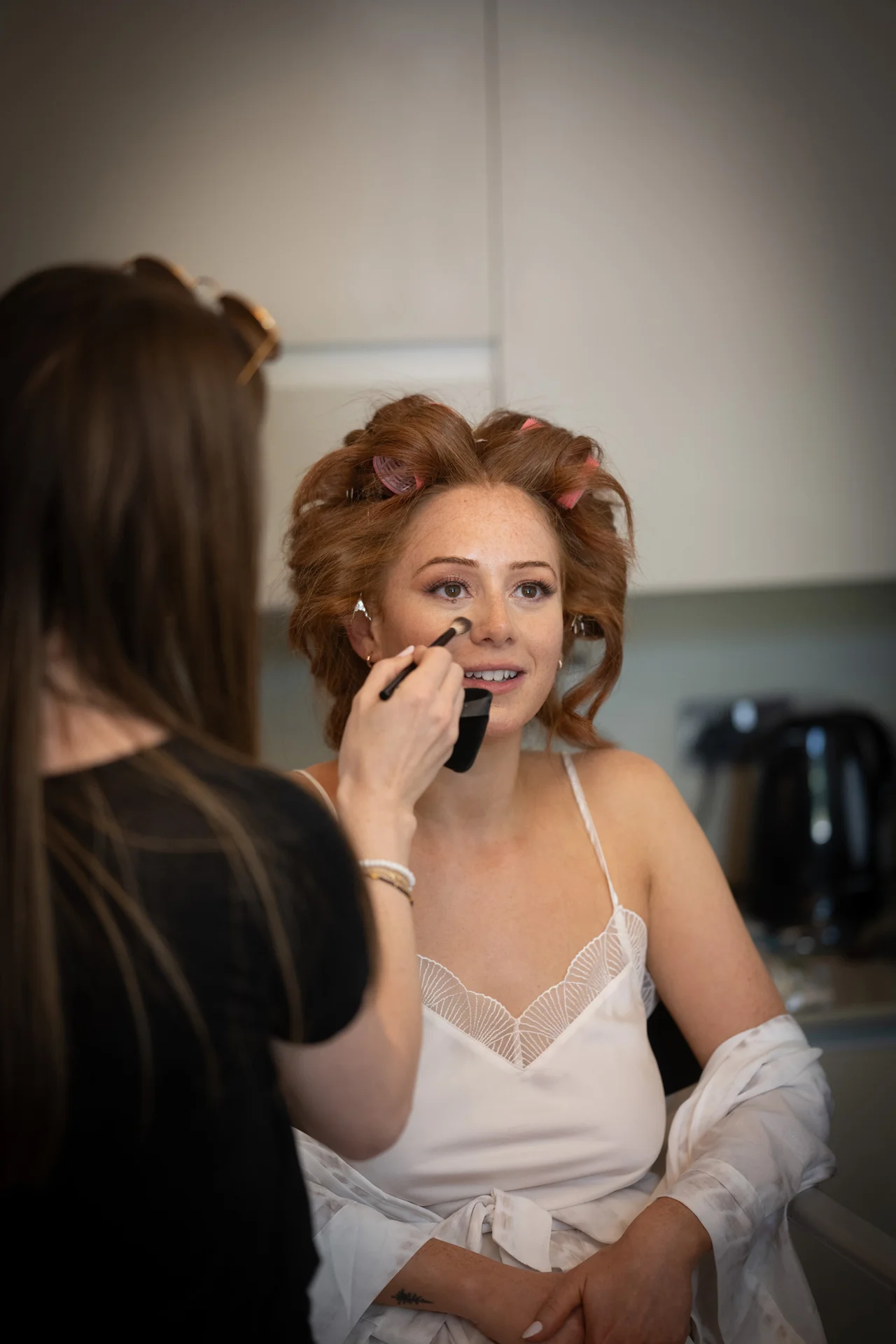 A makeup artist carefully applies finishing touches to a radiant red-haired bride with rollers in her hair as she prepares for her wedding morning.