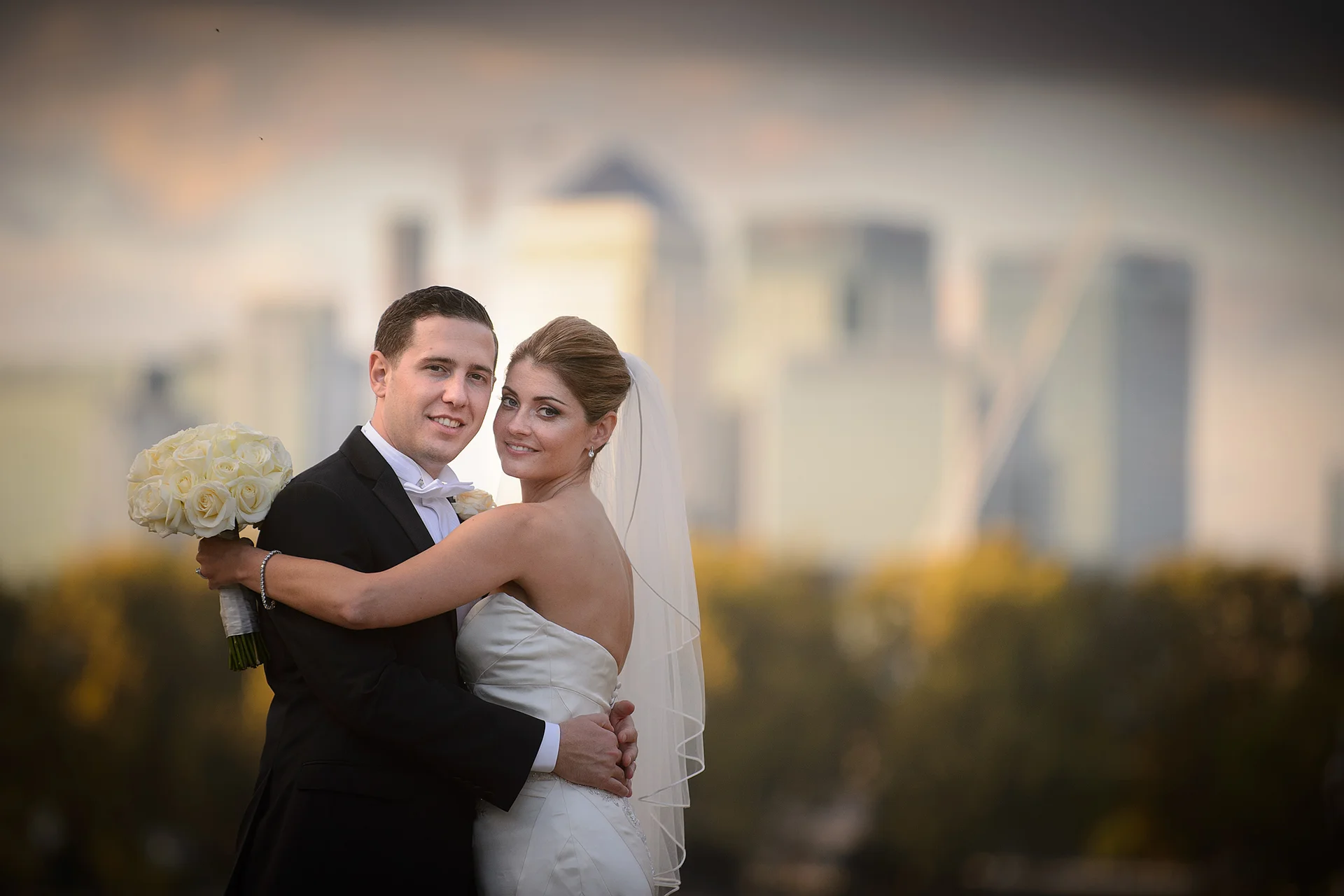 A newlywed couple embraces outdoors against a soft-focus city skyline during twilight, with the bride holding a bouquet of white roses.