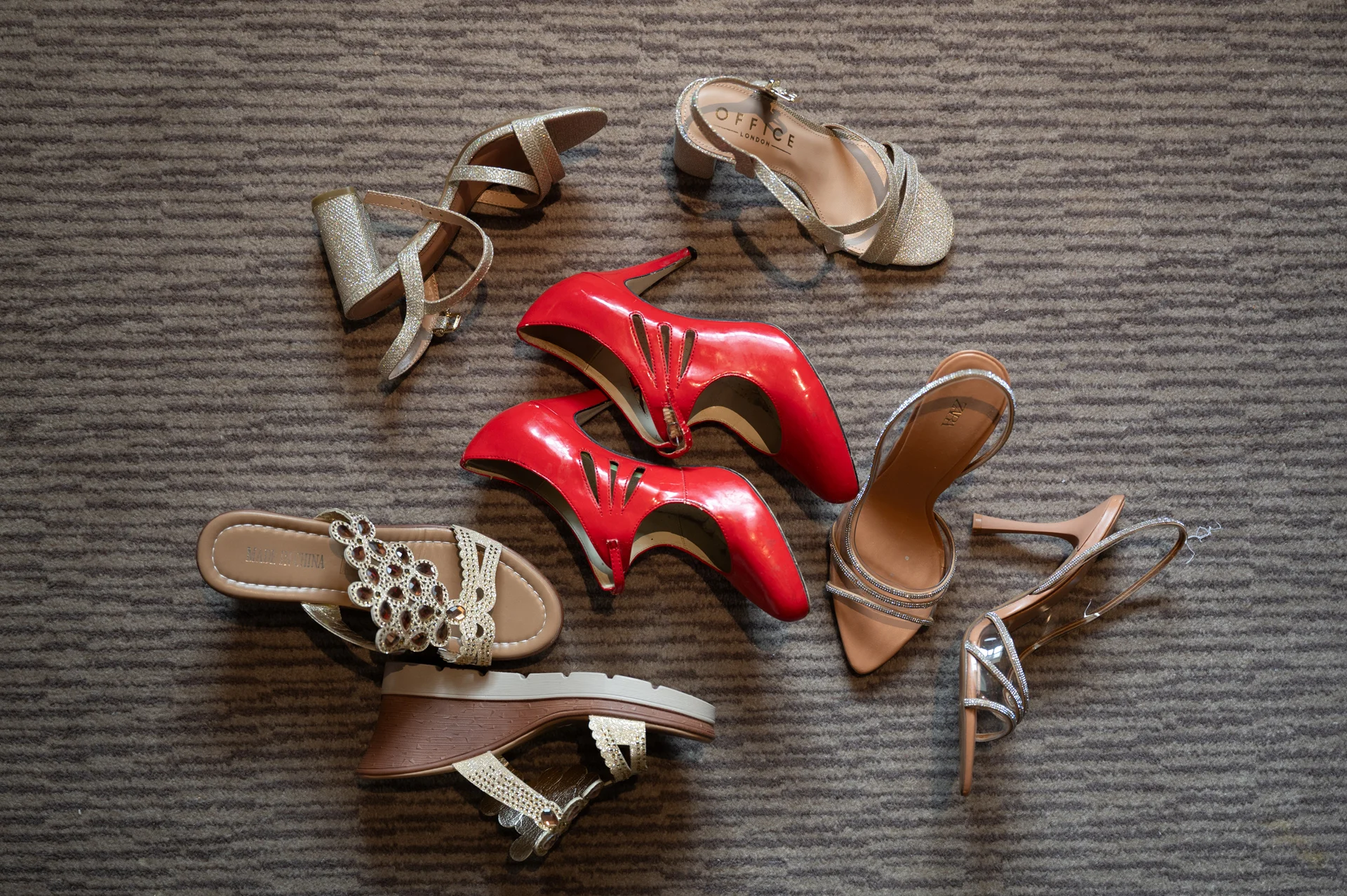 An overhead flat lay captures a stylish collection of bridal shoes, featuring sparkling gold sandals, elegant nude heels, and bold red pumps arranged on a textured grey carpet.