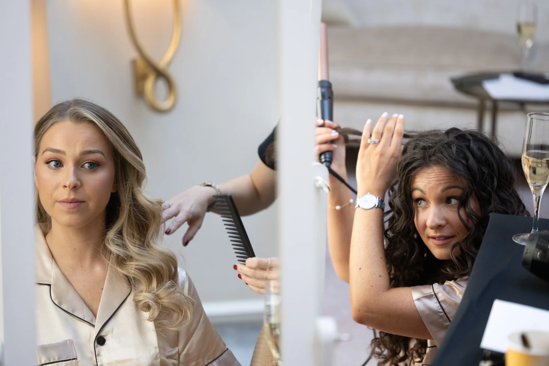 A bride in silk pyjamas has her hair styled during morning bridal preparations while looking into a mirror.
