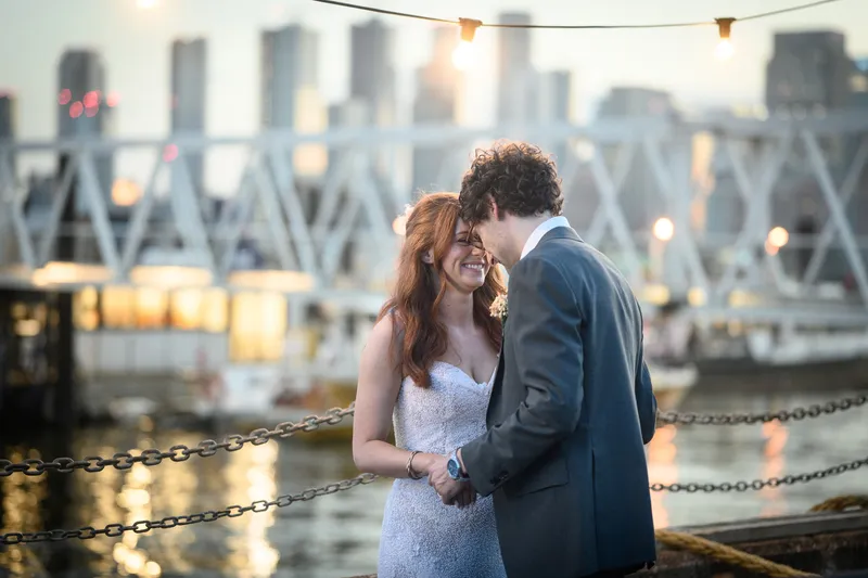 A bride and groom share a tender moment nose to nose against an illuminated urban harbour backdrop at dusk.