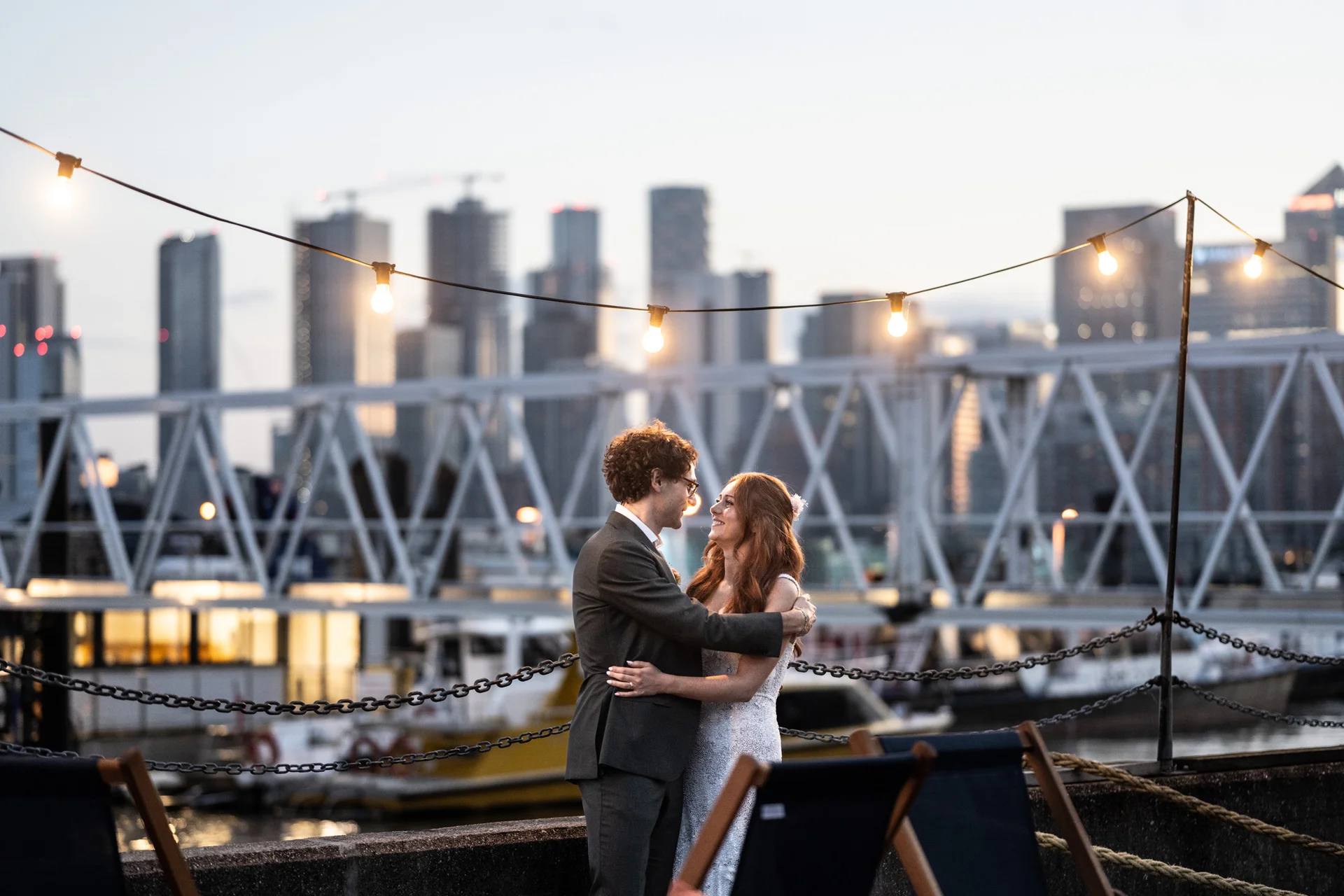 A newlywed couple embraces lovingly on a rooftop terrace at dusk, framed by glowing festoon lights and a striking London city skyline backdrop.