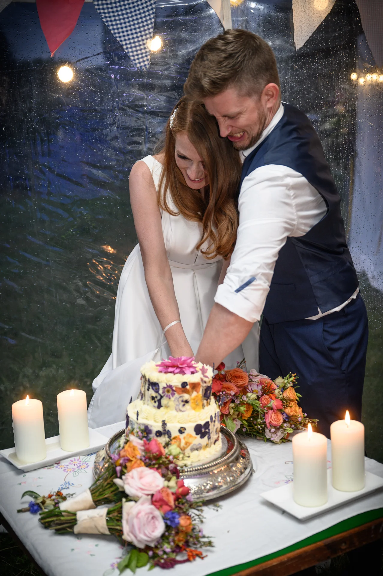 A newlywed couple laughing together while cutting a two-tier pressed flower wedding cake inside a candlelit marquee on a rainy evening.