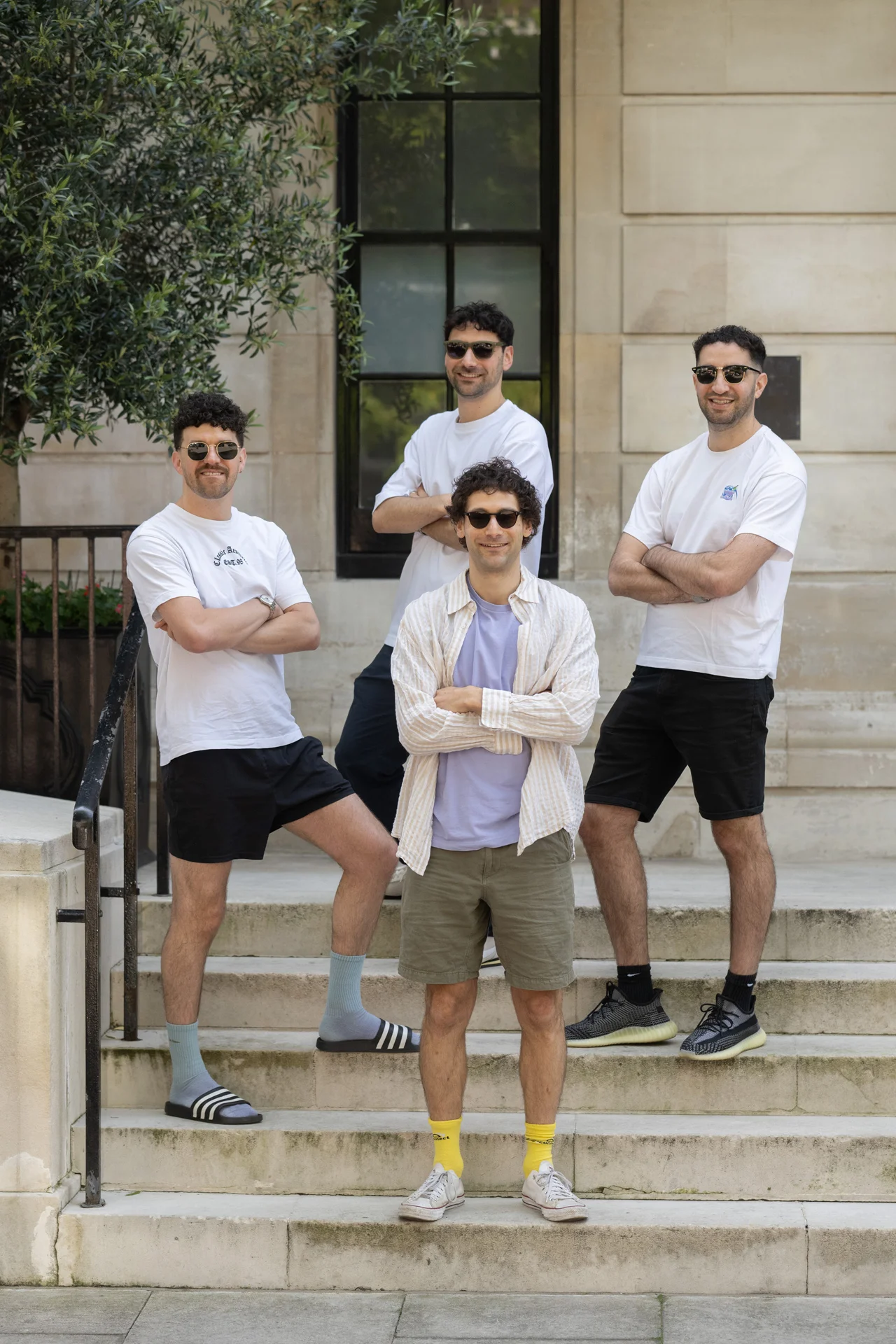 Four male guests pose casually with sunglasses and folded arms on stone steps outside an urban wedding venue.
