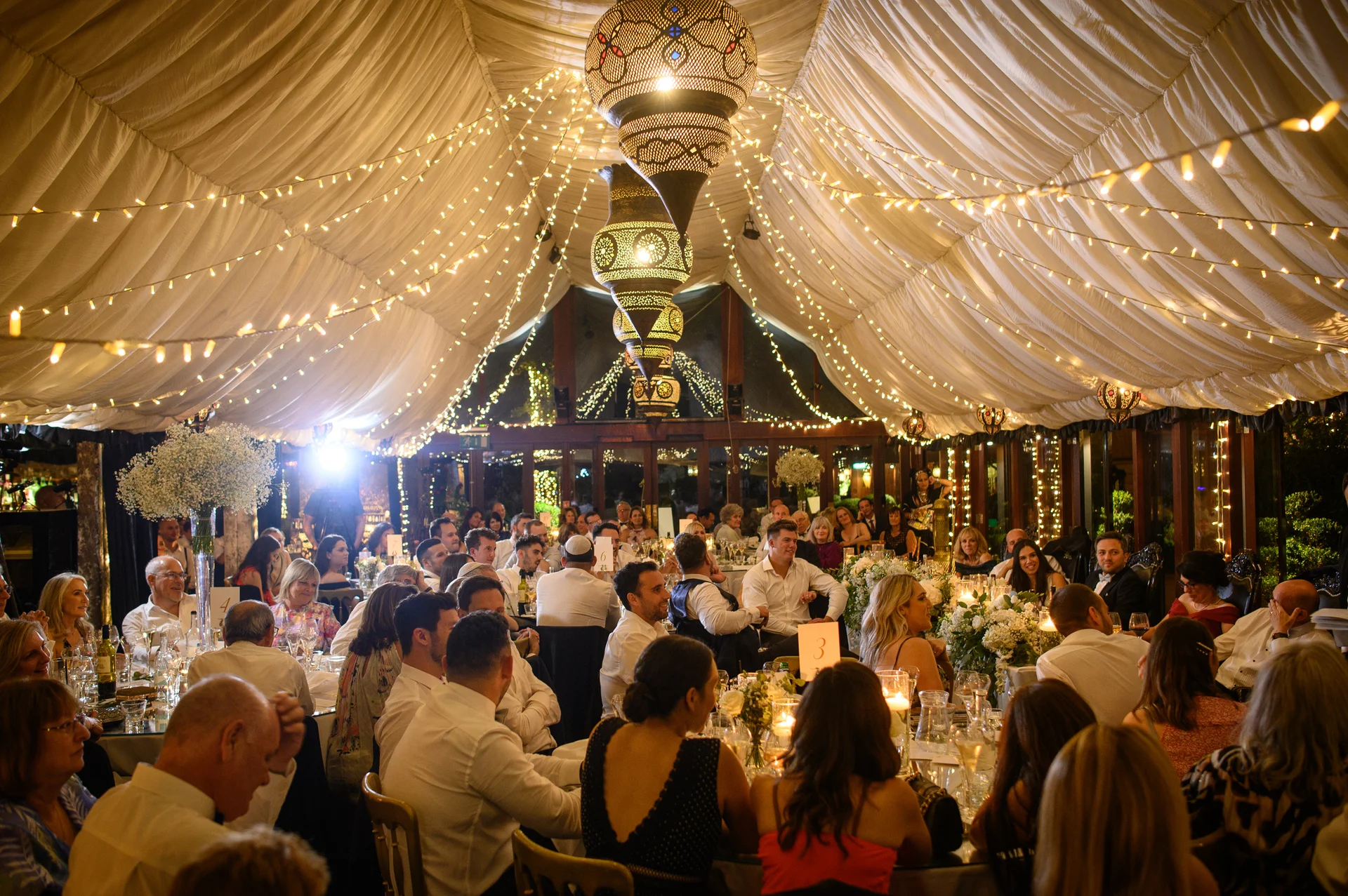 Guests enjoy a wedding breakfast inside a marquee draped in fabric and illuminated by a dazzling canopy of fairy lights and hanging lanterns.
