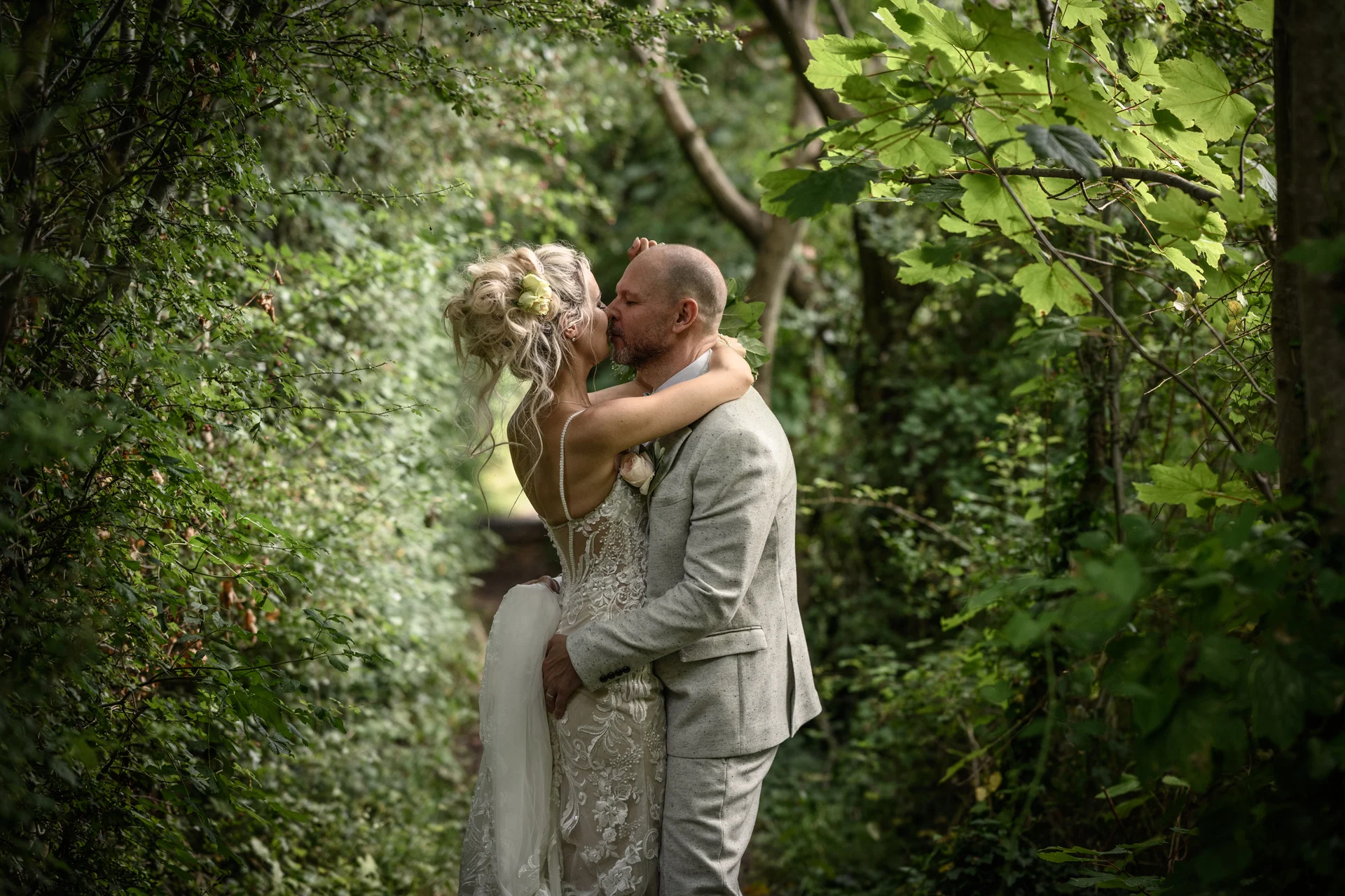 A newlywed couple share a tender kiss while surrounded by lush green woodland foliage and dappled sunlight.