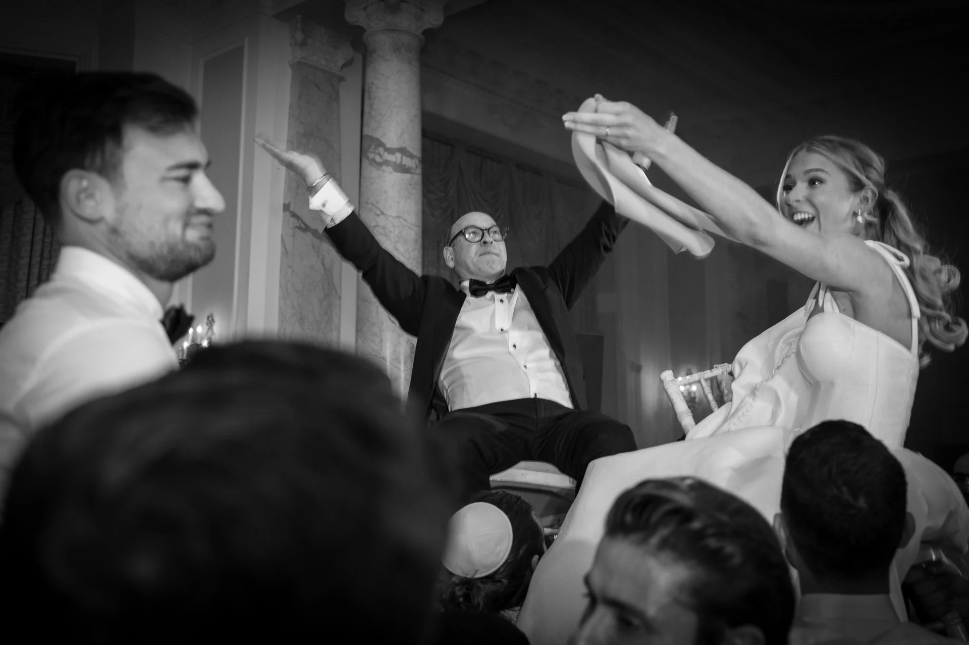 A joyful black and white image capturing wedding guests lifting the couple high in chairs during an energetic hora dance celebration.