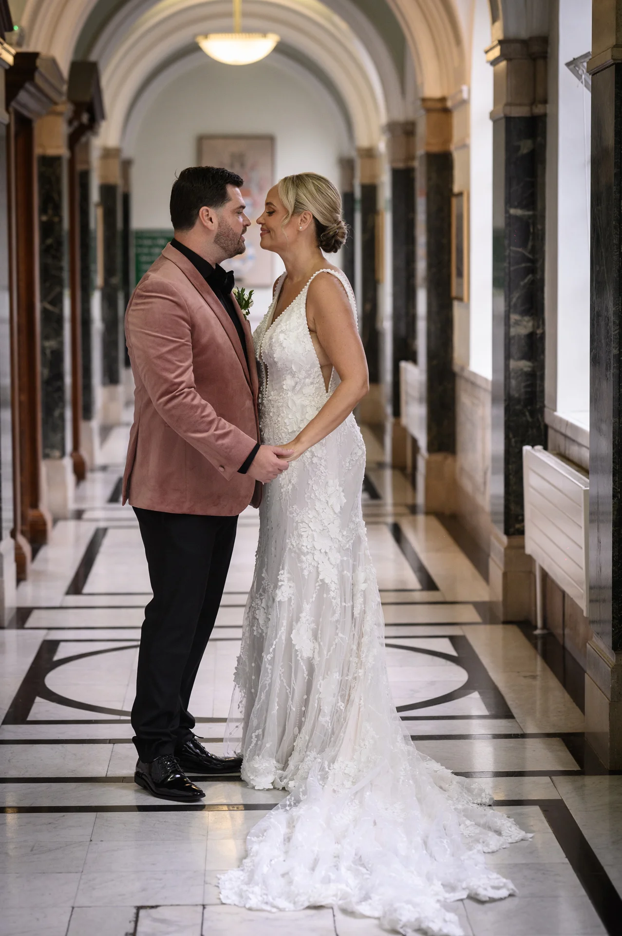 A newlywed couple gazes lovingly at each other nose to nose inside a grand historic hallway with geometric marble flooring.