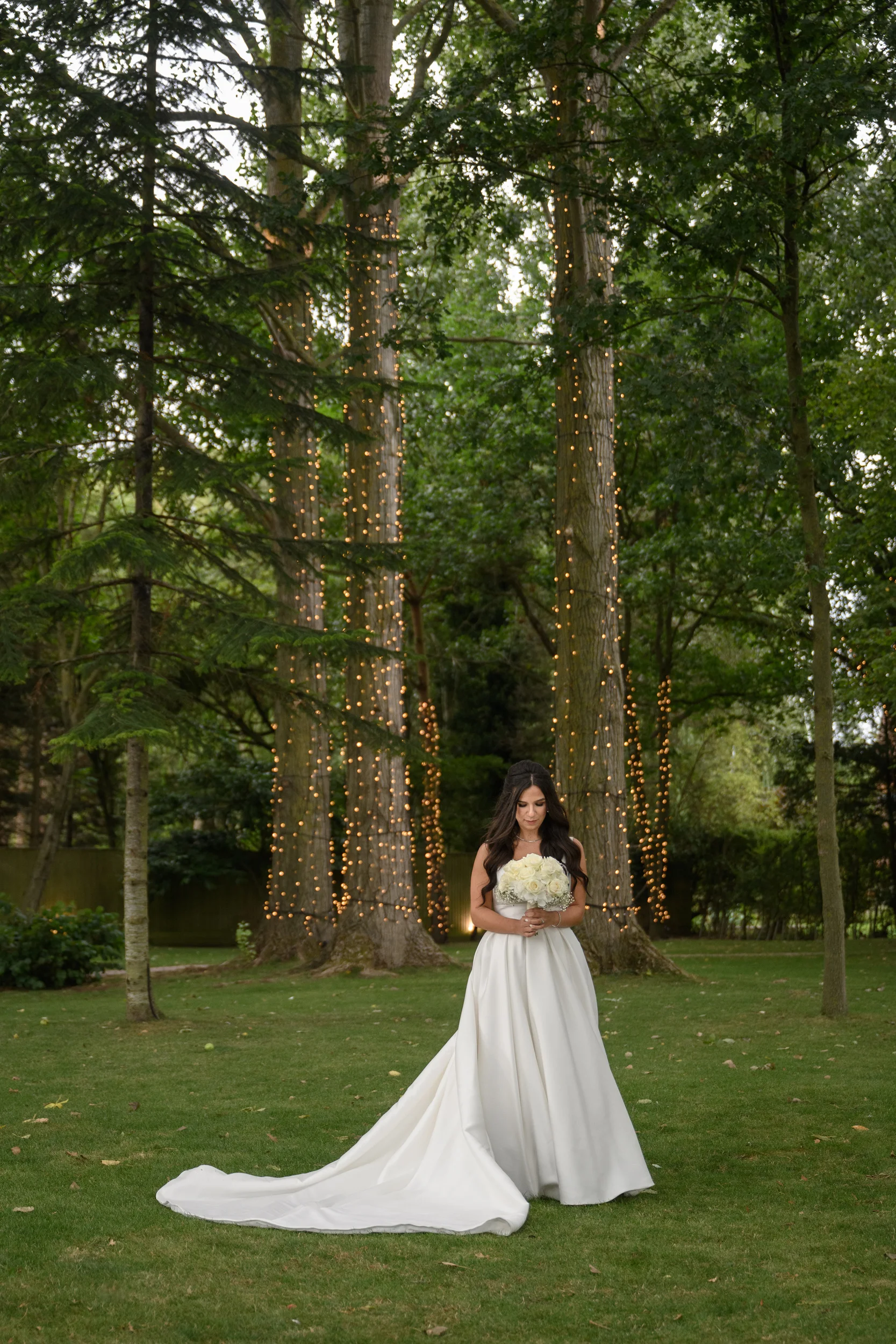 A stunning bride stands in a lush woodland setting with a fairy light canopy wrapped around the trees, holding a classic bouquet of white roses.