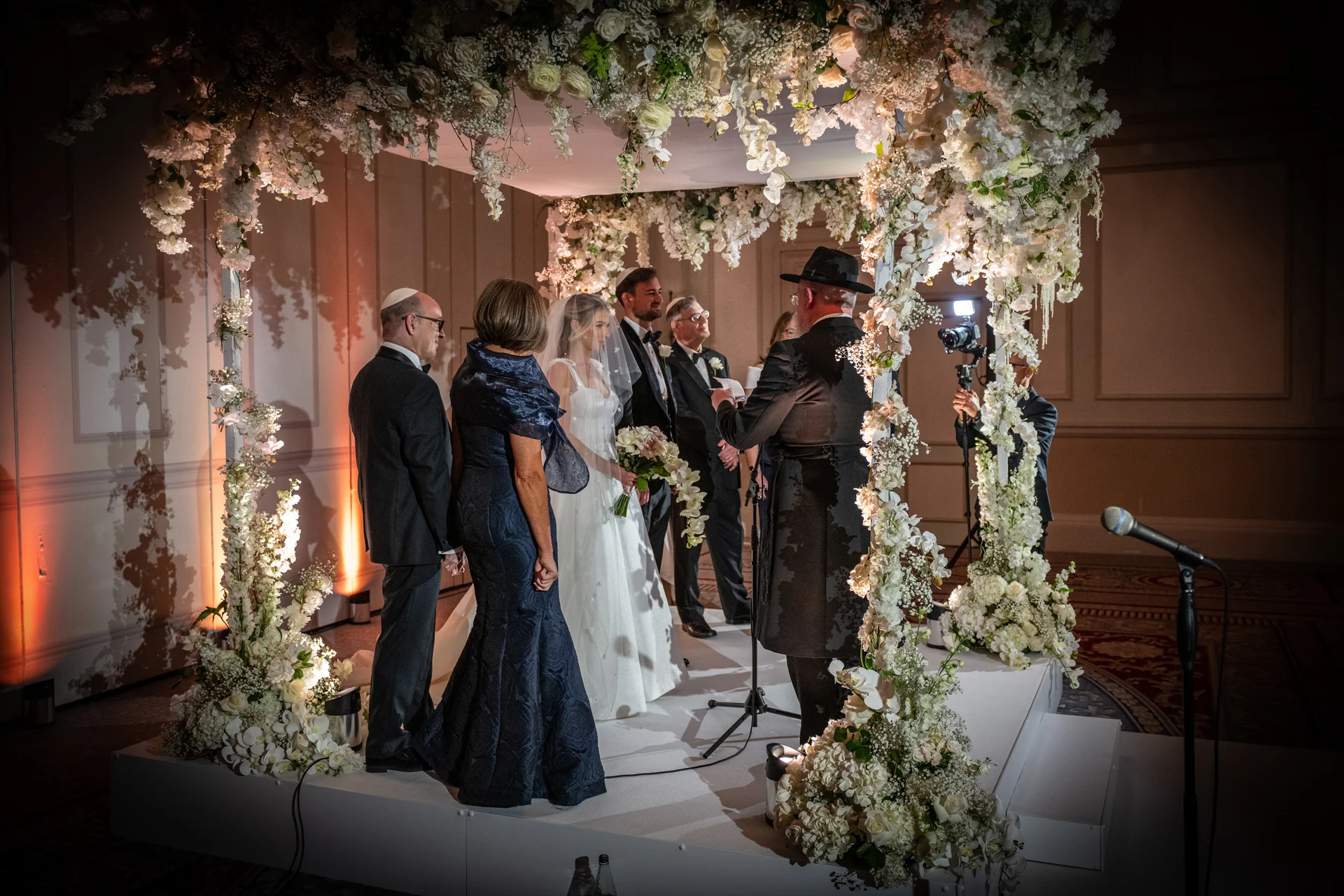 A Jewish wedding ceremony takes place beneath an elaborate white floral chuppah with family members and officiants gathered around the bride and groom.