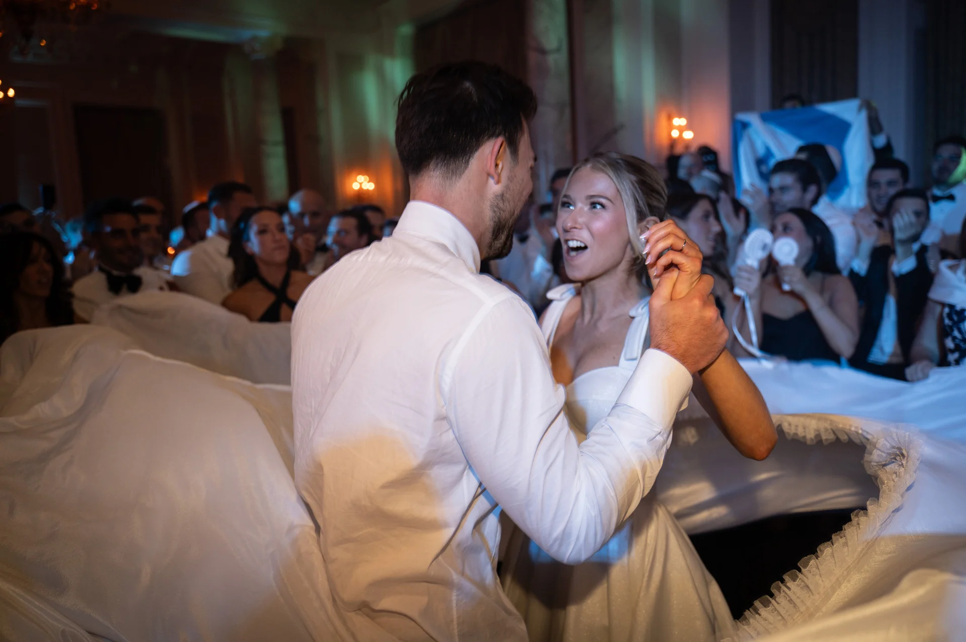 A newlywed couple shares an energetic first dance surrounded by cheering wedding guests in an elegant ballroom.