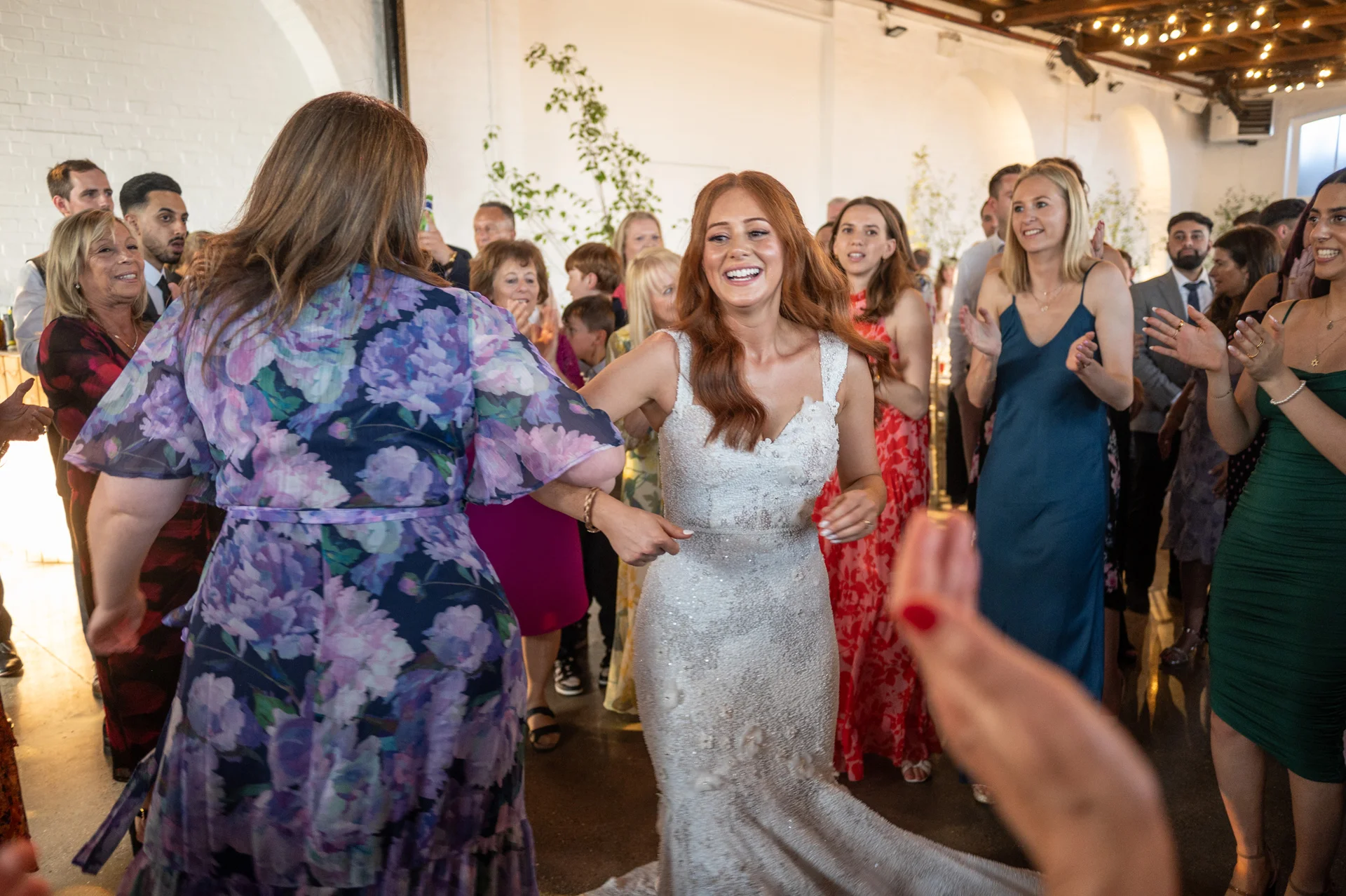 A joyful bride with long red hair smiles radiantly as she dances on a lively reception dance floor surrounded by cheering guests.