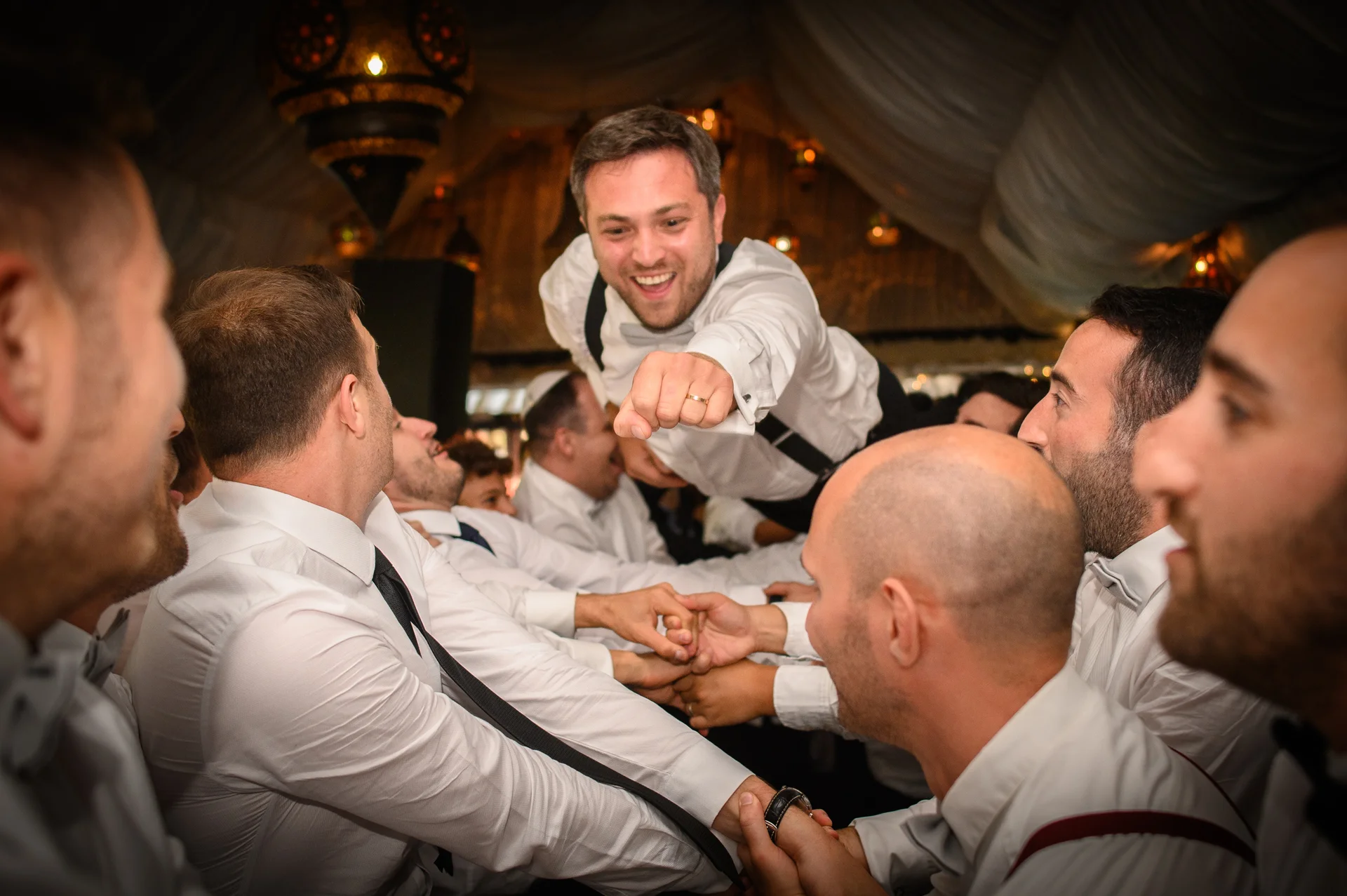 A joyful groom is lifted into the air by cheering groomsmen during the lively reception celebration inside a decorated marquee.