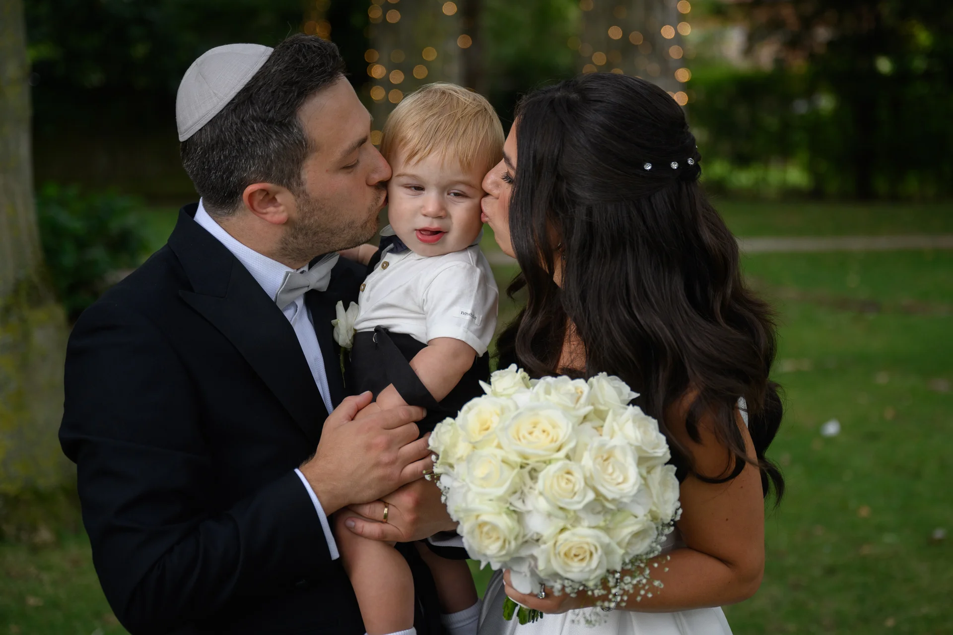 A bride and groom lovingly kiss their young page boy on the cheeks while holding him in an outdoor garden setting.