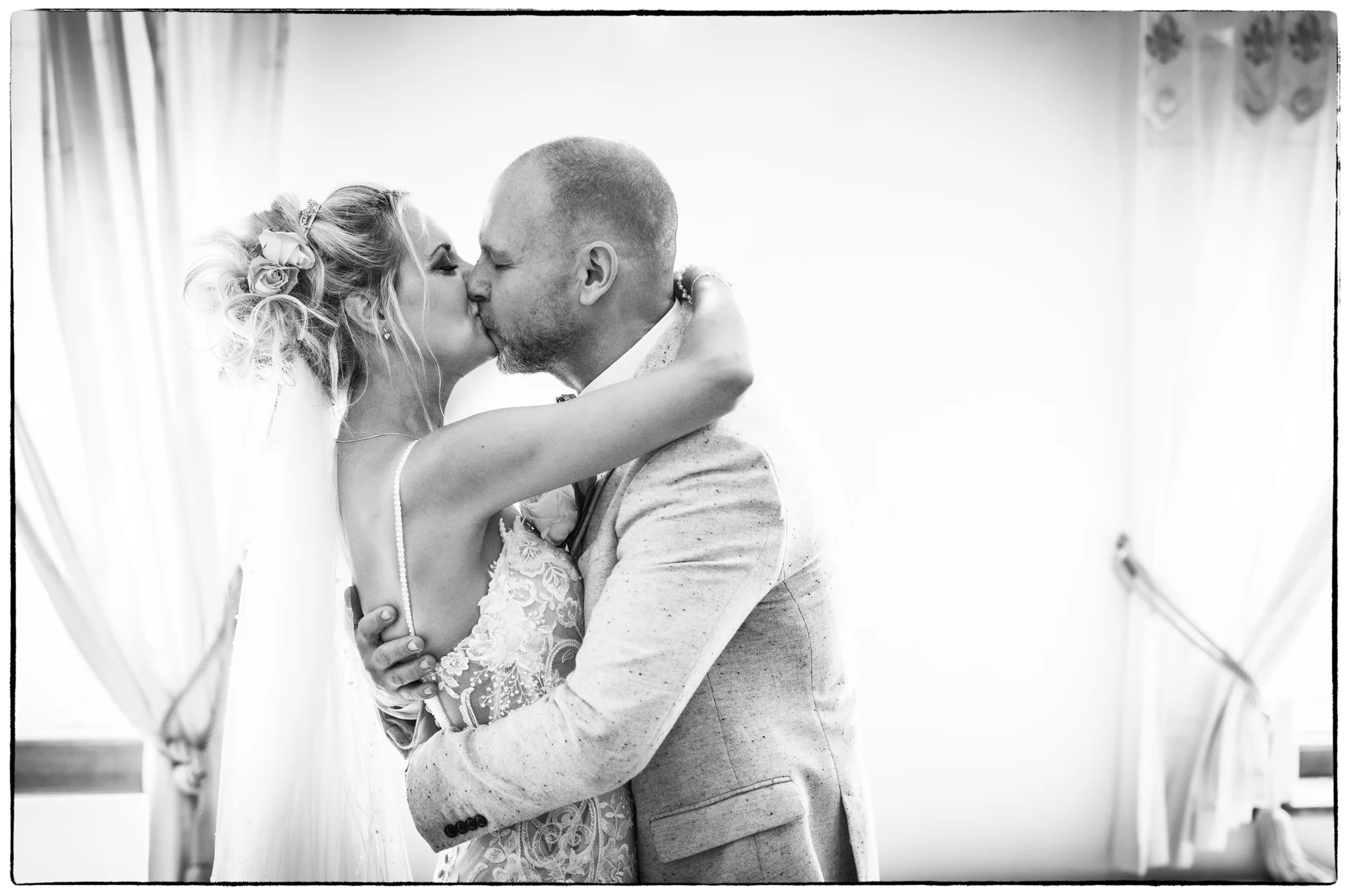 A bride in a lace gown and groom in a patterned jacket share a tender kiss in silhouette against a bright window.