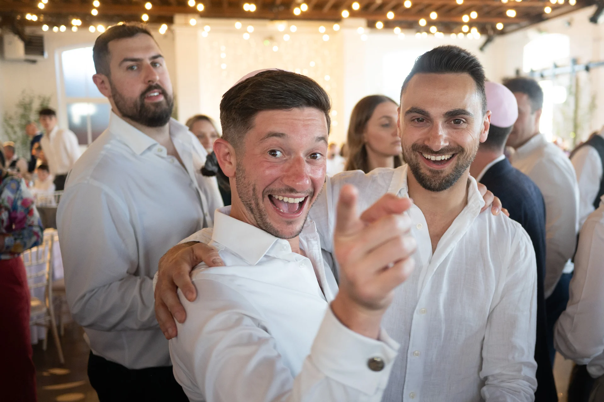 A joyful couple embraces closely inside a bright reception venue featuring exposed wooden rafters and overhead string lights, laughing happily as guests mingle around them.