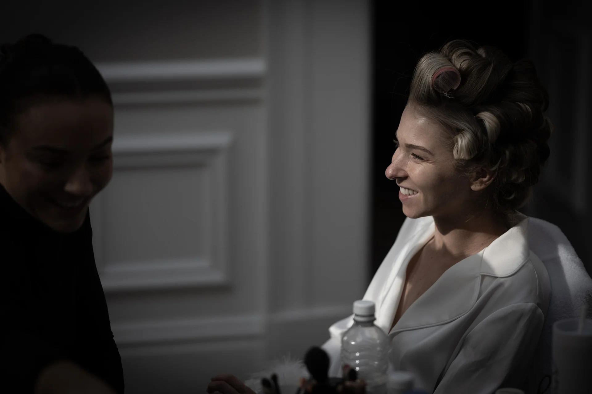 A smiling bride wearing hair rollers and a white robe laughs during wedding morning preparations while sitting near another person.