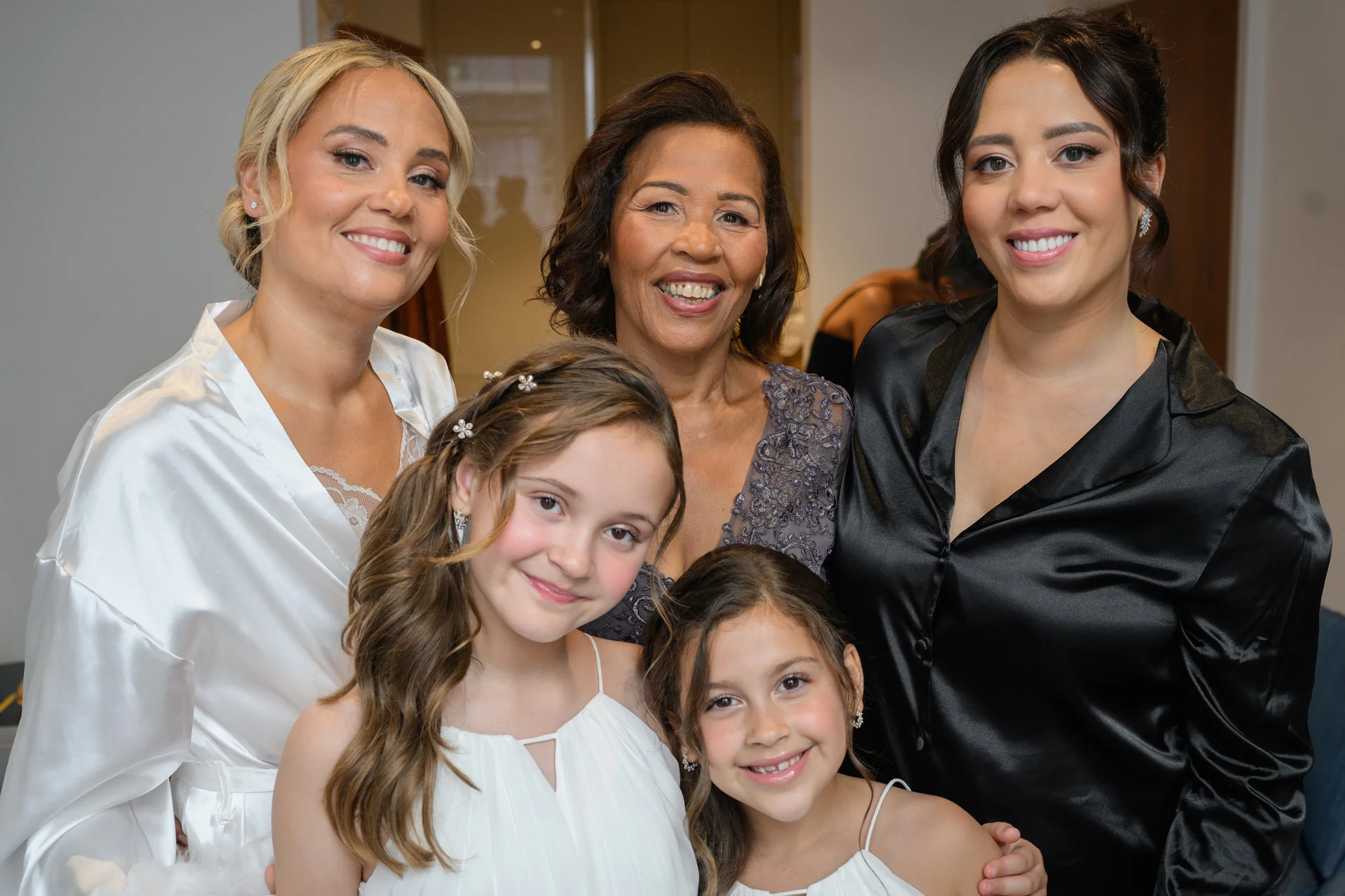 A joyful group of family members including a mother, bridesmaids, and young flower girls smiling together indoors while getting ready for the wedding.
