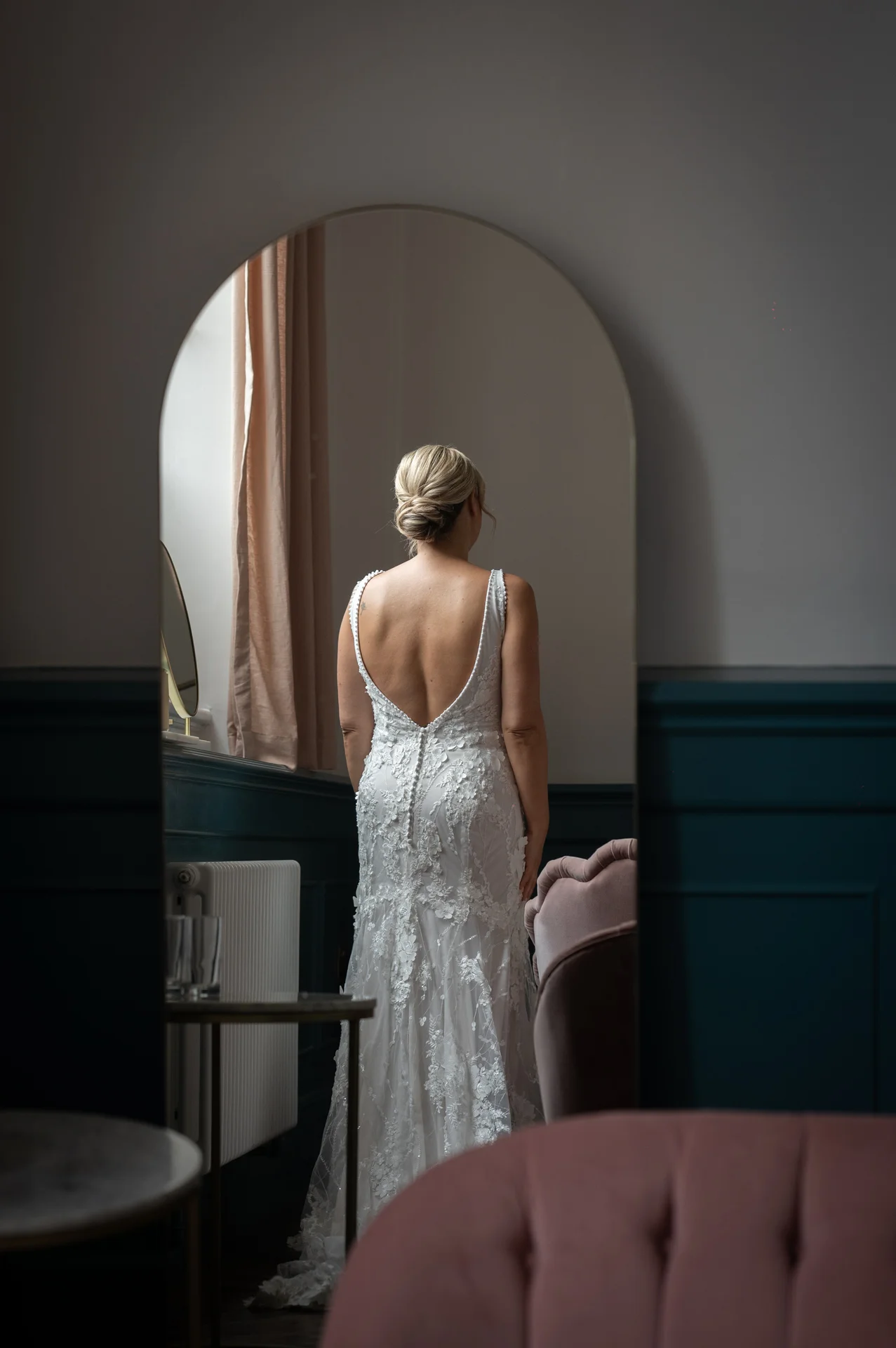 A bride stands facing an arched mirror, showcasing the stunning lace details and open back of her wedding gown during morning preparations.