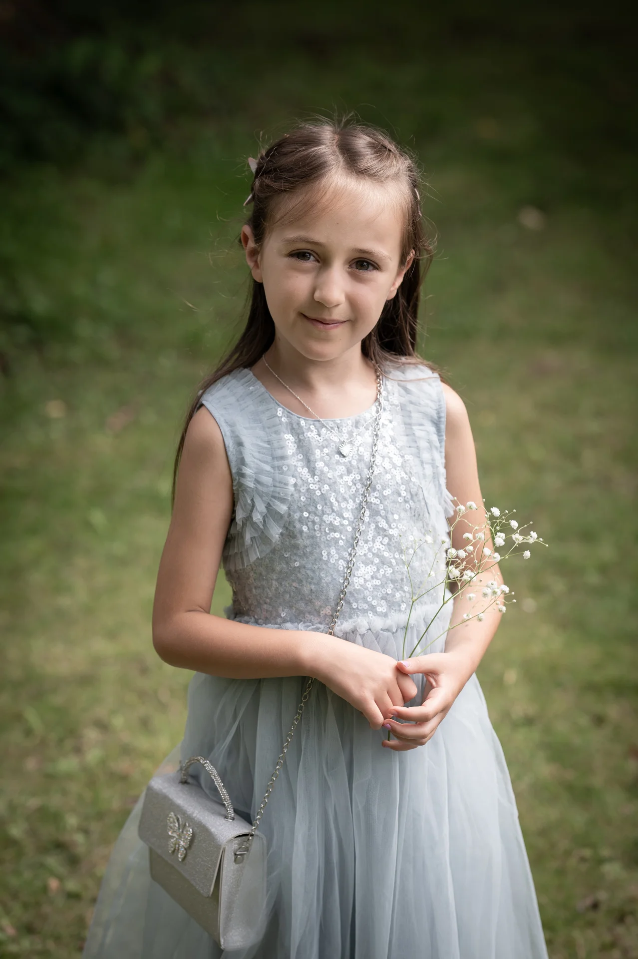 A young flower girl stands outdoors in a soft blue tulle dress, holding a delicate sprig of gypsophila and a small silver handbag.