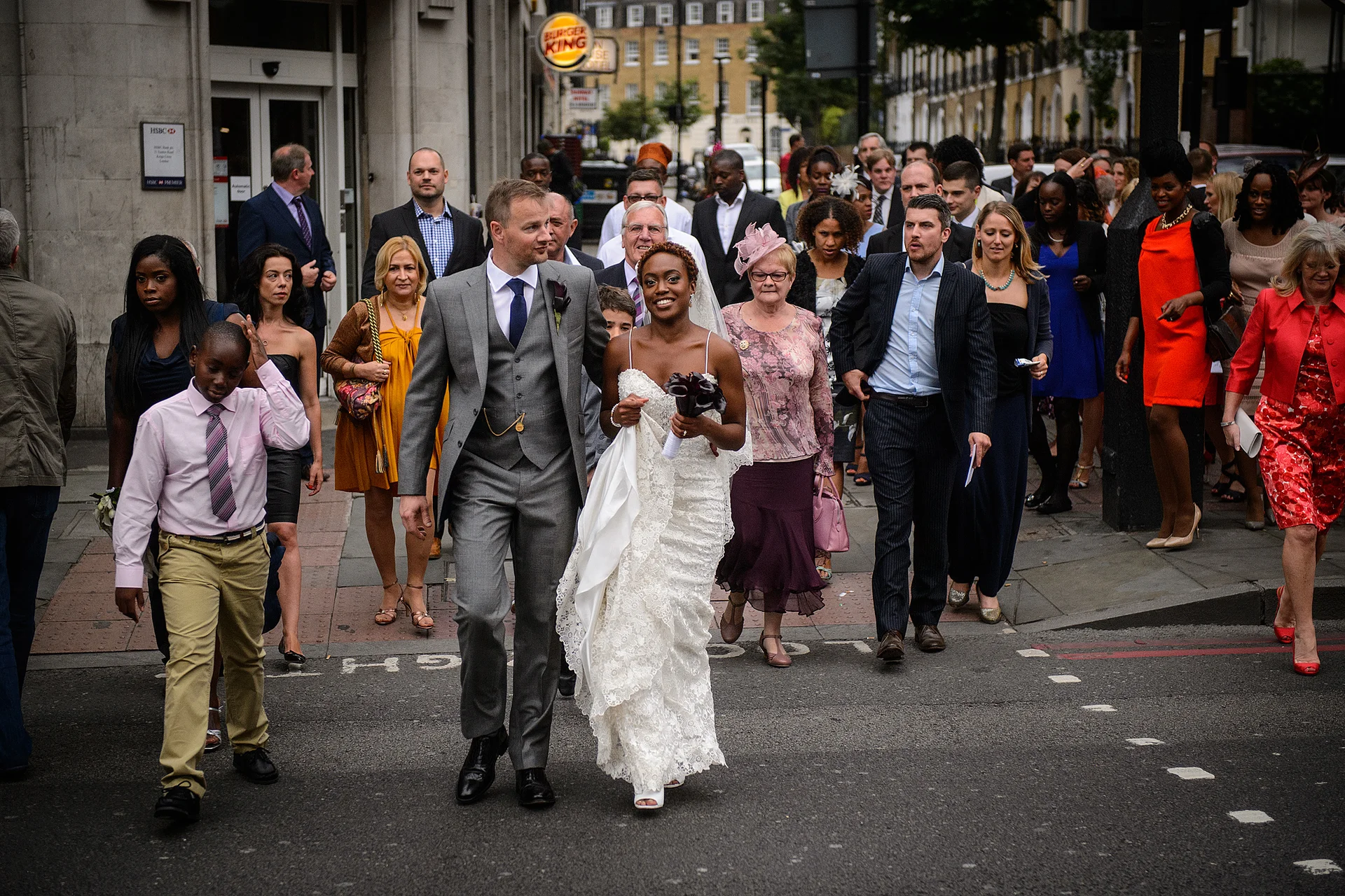 A smiling bride and groom walk hand in hand across a London street followed by a large crowd of wedding guests.