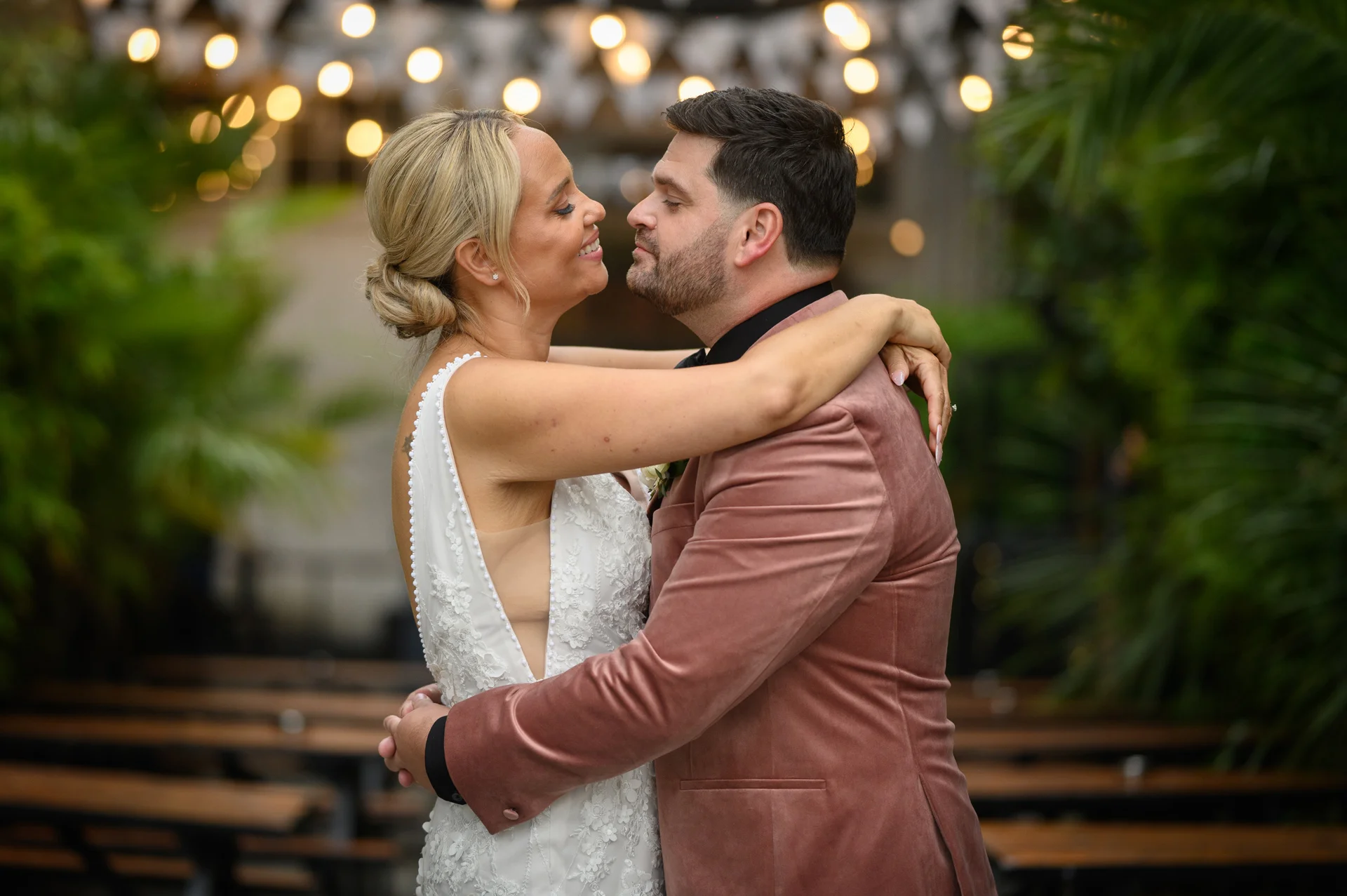 A newlywed couple embraces lovingly outdoors under a canopy of warm festoon lights, the bride in a lace gown and the groom in a stylish pink velvet jacket.