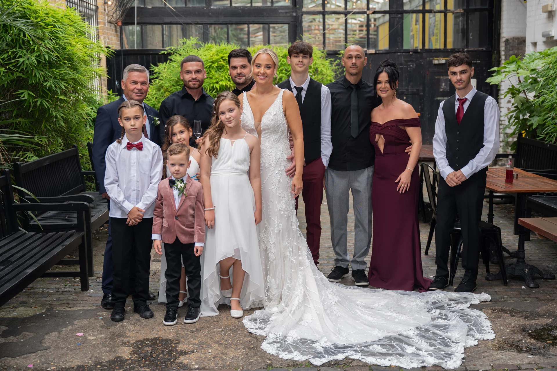 A newlywed bride in a plunging floral lace gown poses with her extended family and bridal party in a cobbled industrial courtyard.