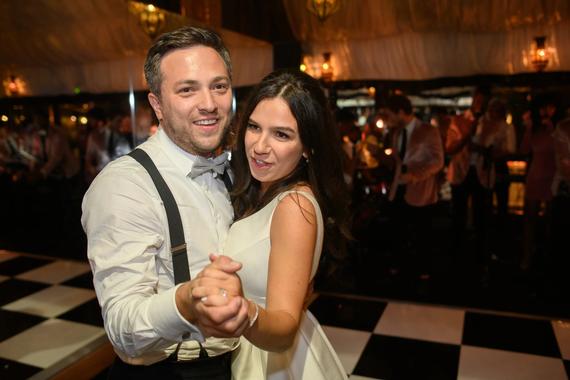 A smiling newlywed couple shares a joyful first dance on a black and white checkerboard dance floor during their evening reception.