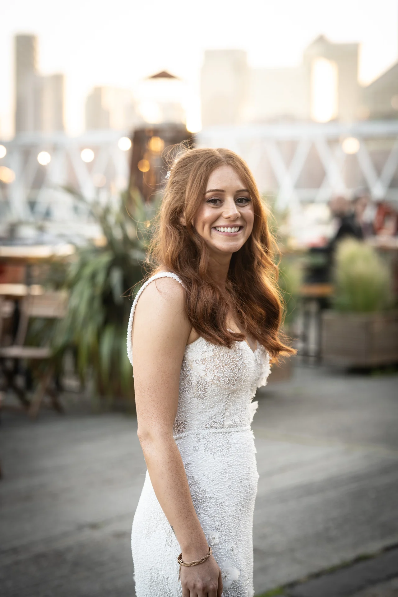 A stunning portrait of a smiling bride with long red hair wearing a beaded gown at an urban rooftop wedding venue.