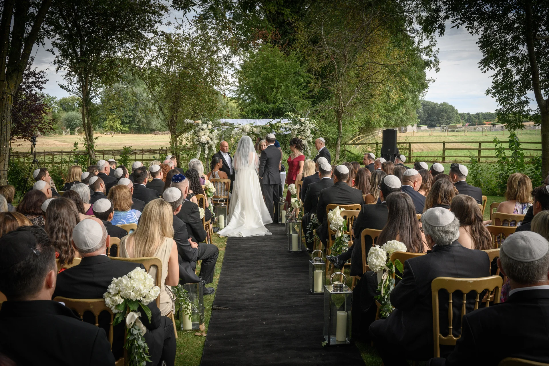 An outdoor Jewish wedding ceremony takes place beneath a floral-adorned chuppah, with guests seated on either side of a black aisle runner.