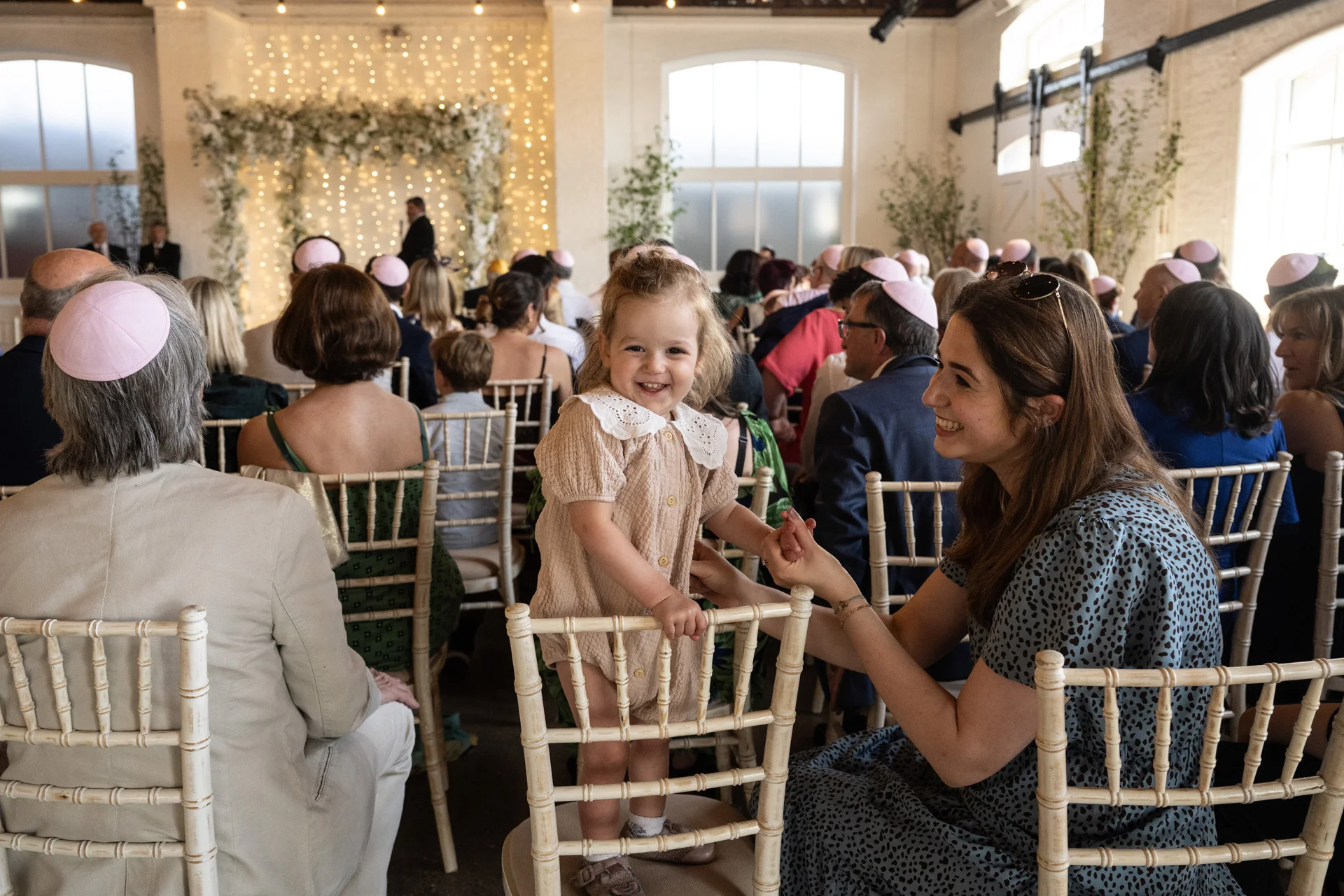 A smiling toddler stands on a white chiavari chair during a light-filled indoor wedding ceremony, holding hands with a seated guest while guests look on.