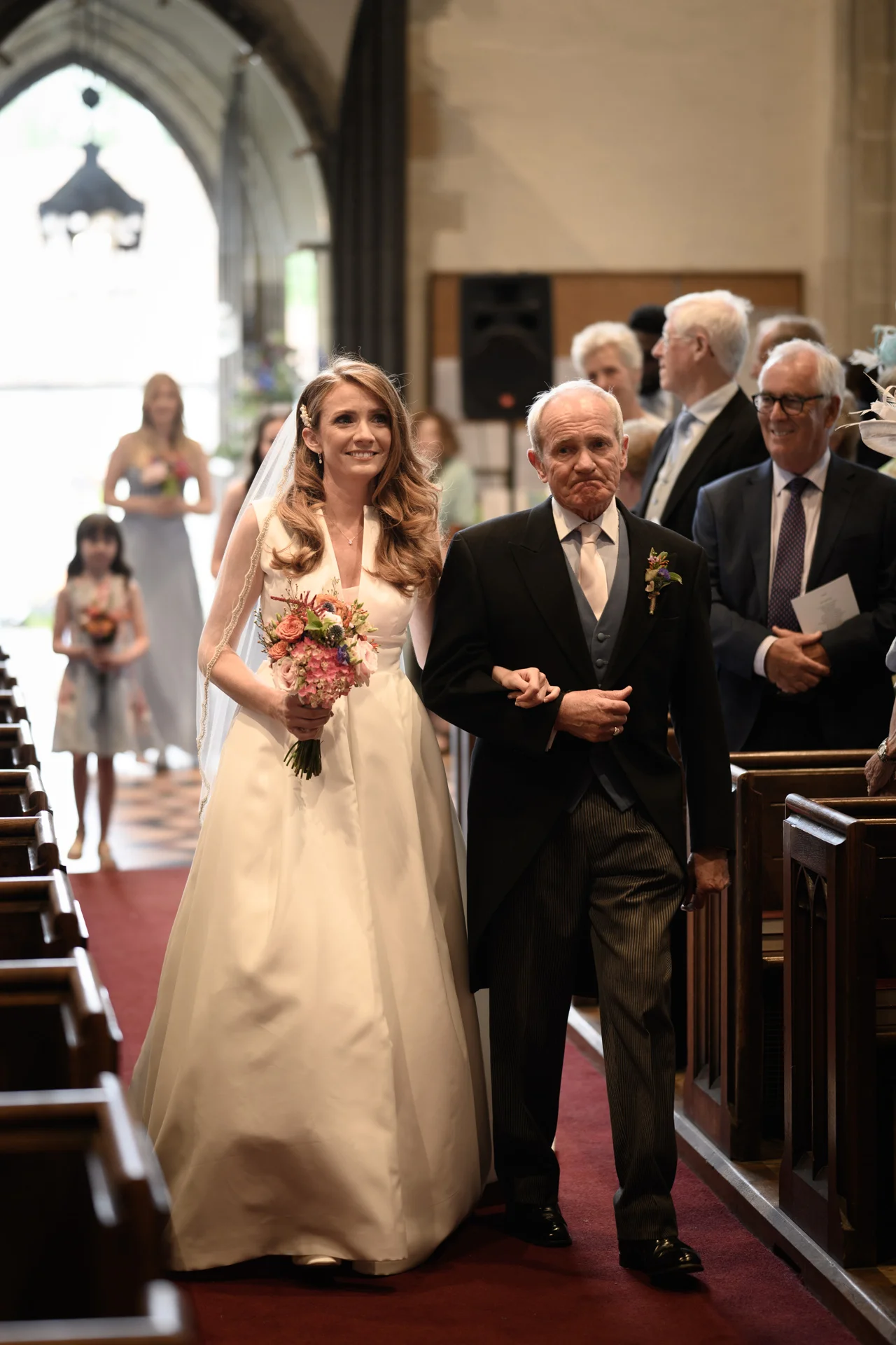 A smiling bride holding a colourful bouquet walks down the church aisle arm in arm with her father in a morning suit, flanked by wooden pews and watching guests.