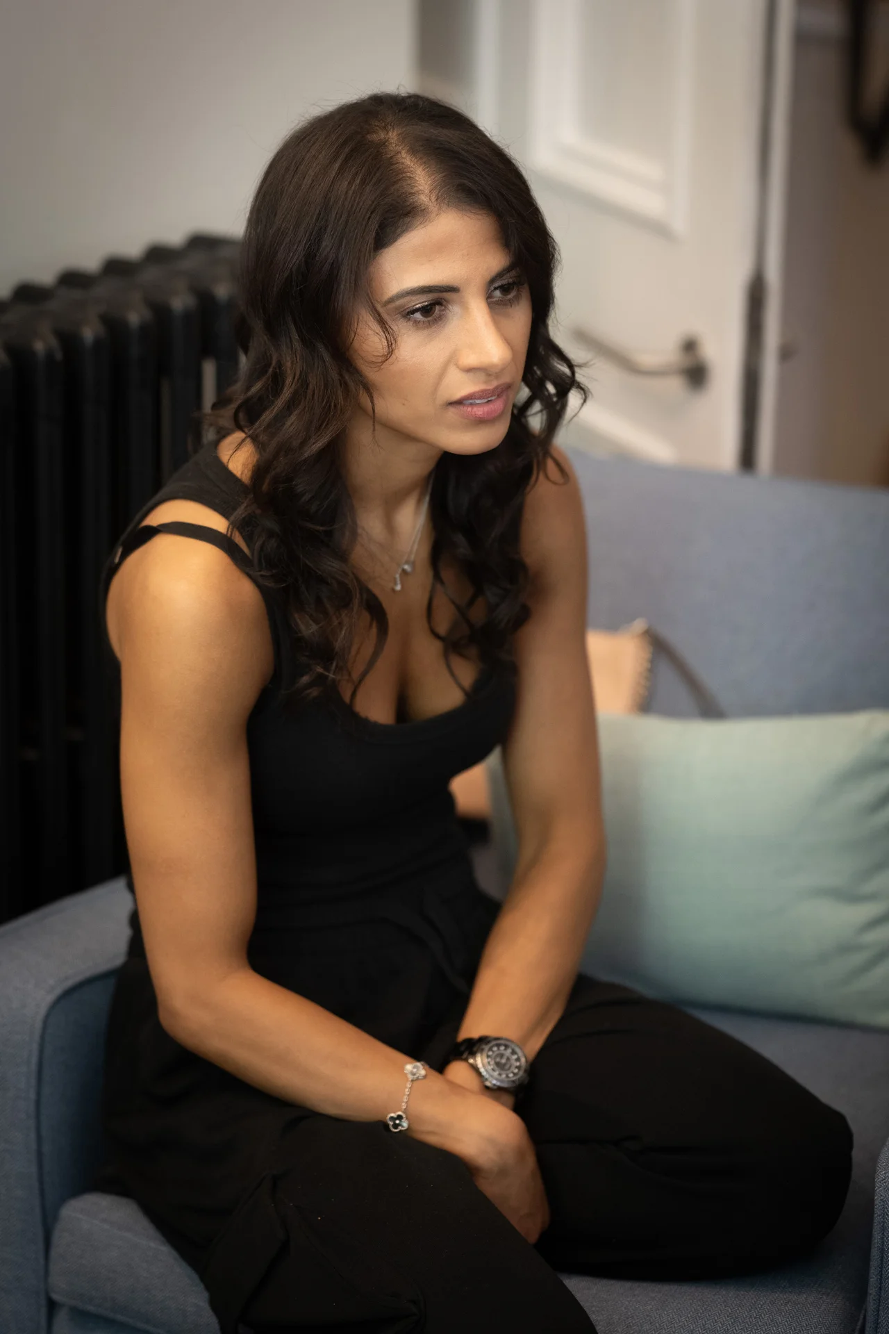 A woman with dark wavy hair and a black outfit sits thoughtfully on a blue sofa during wedding morning preparations.