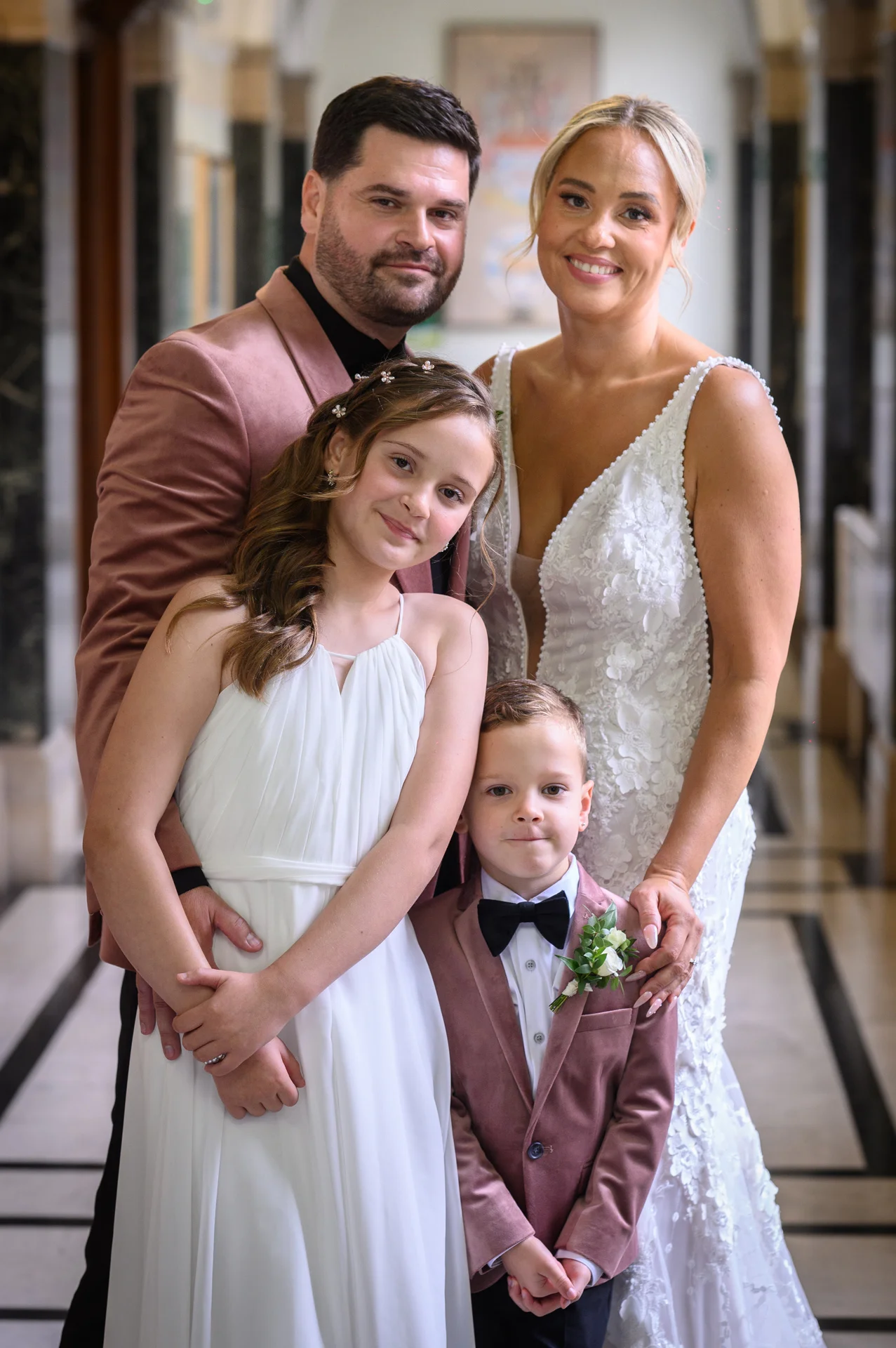 A smiling family of four poses together indoors, with the bride wearing a beaded gown and the groom in a pink velvet jacket alongside their young children.