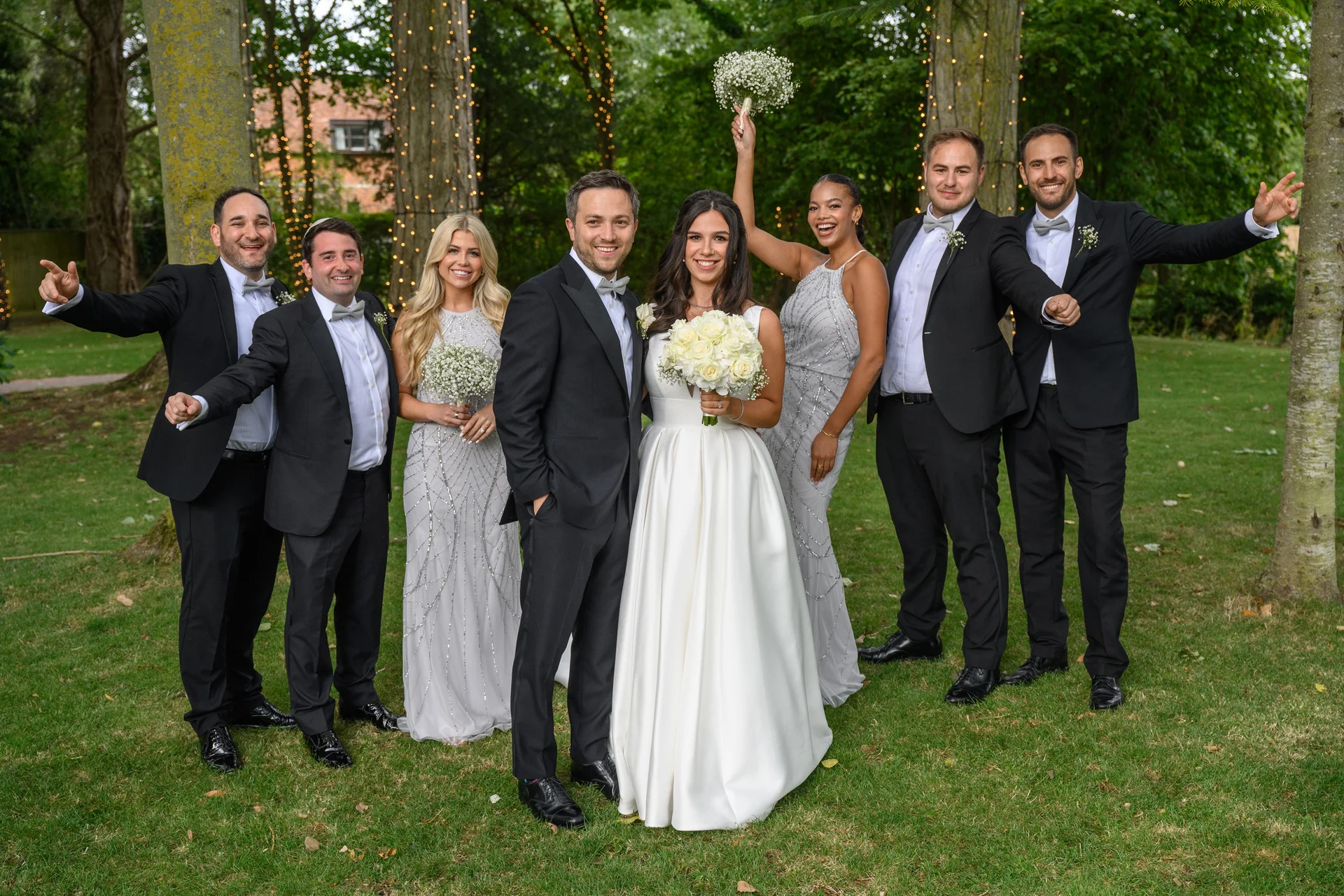 The smiling bride and groom stand proudly at the center of a joyful wedding party in a lush garden setting illuminated by fairy lights.