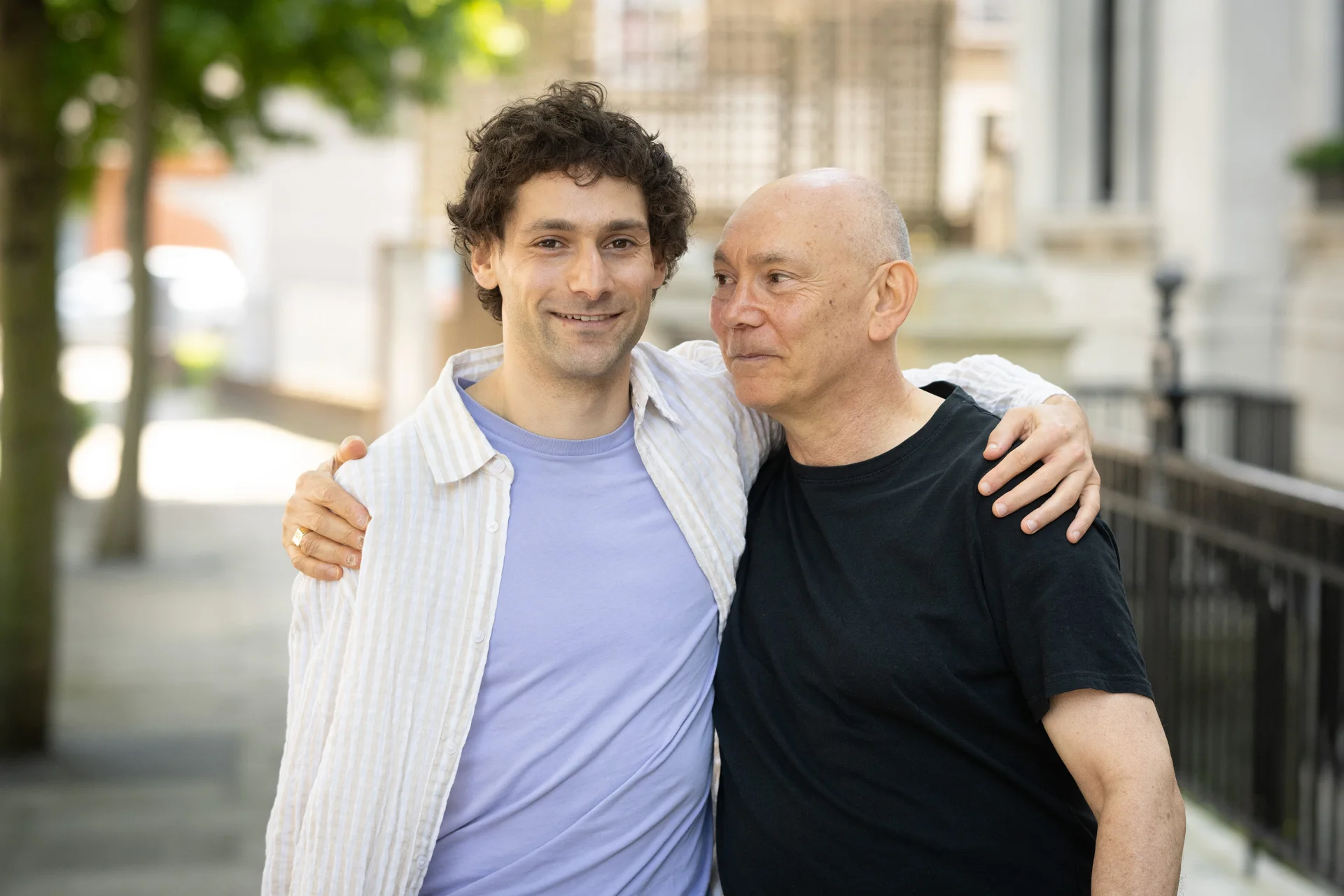 Two grooms embrace affectionately outdoors during a relaxed city wedding celebration in London, smiling warmly as they stand side by side.