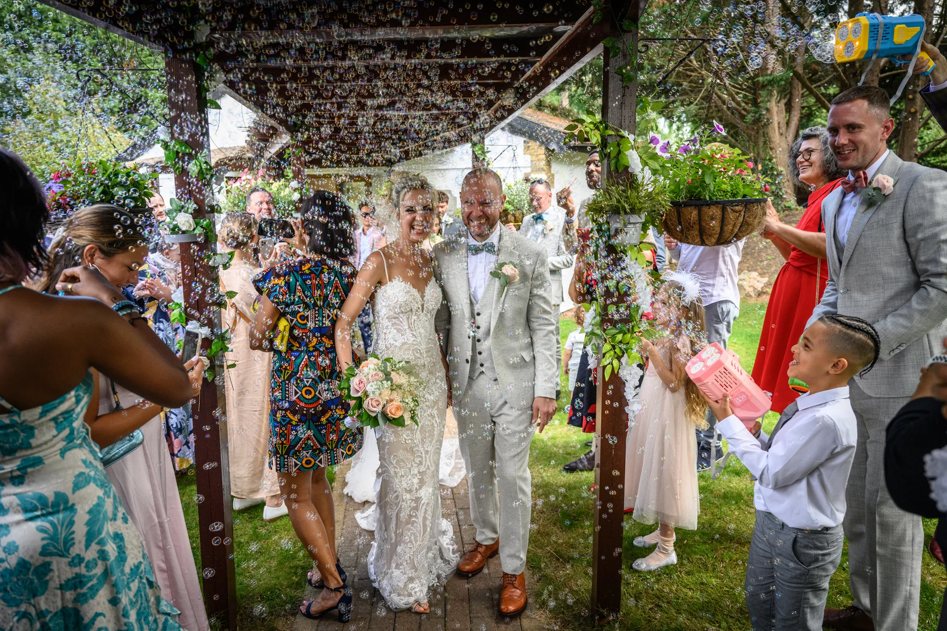 A joyful bride and groom walk through an outdoor wooden pergola as cheering guests blow bubbles during their wedding celebration.