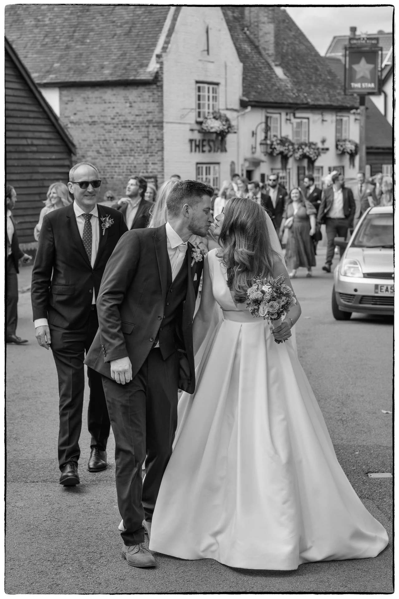 A bride and groom share a sweet kiss while walking down a village street past a traditional British pub, accompanied by wedding guests and a classic car.