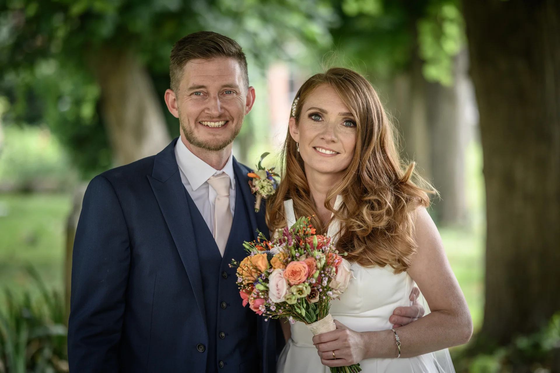 A smiling bride and groom pose together outdoors amidst lush green trees, with the bride holding a colourful hand-tied bouquet.