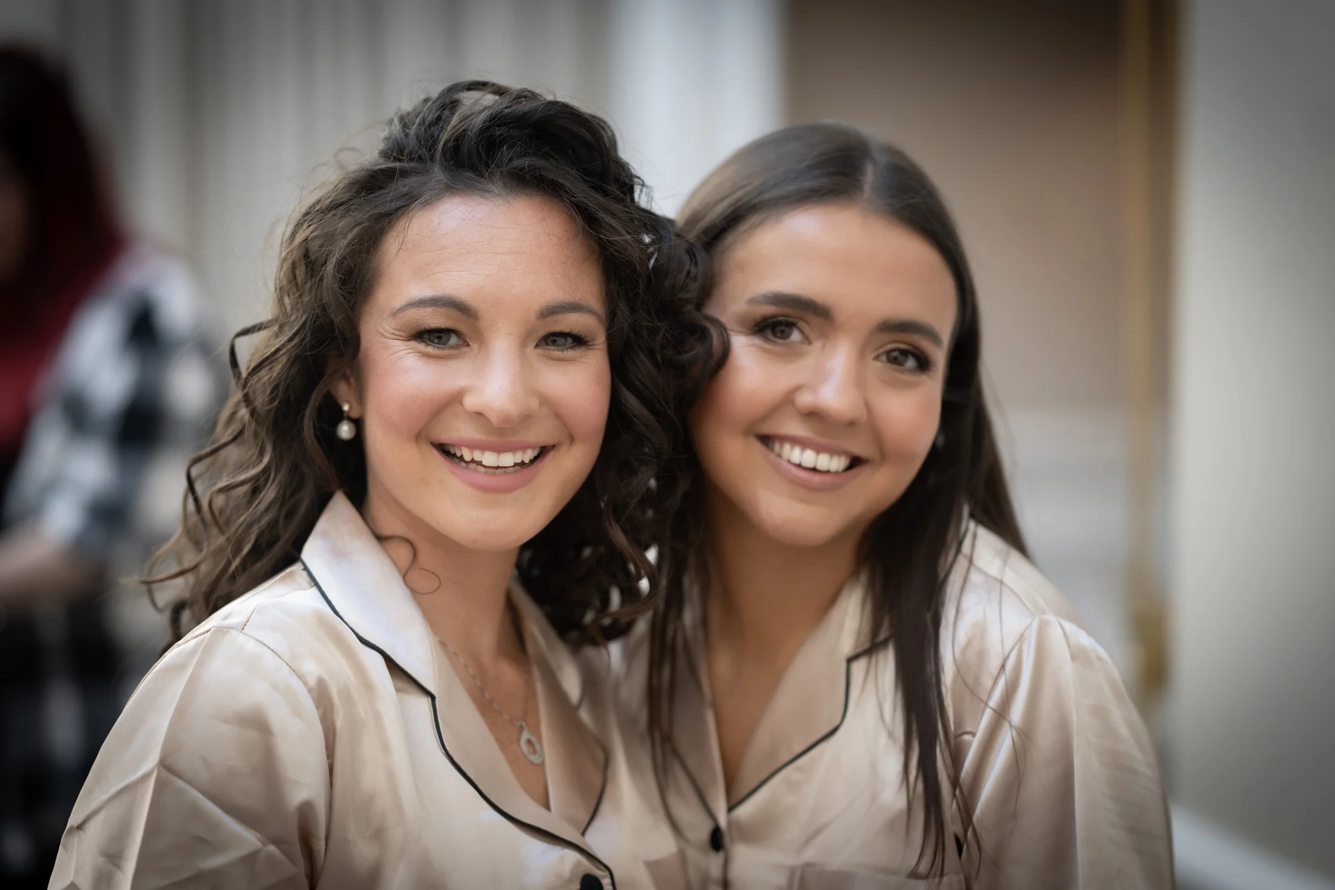 Two smiling women pose closely together indoors while wearing matching champagne-coloured silk pyjamas during wedding morning preparations.