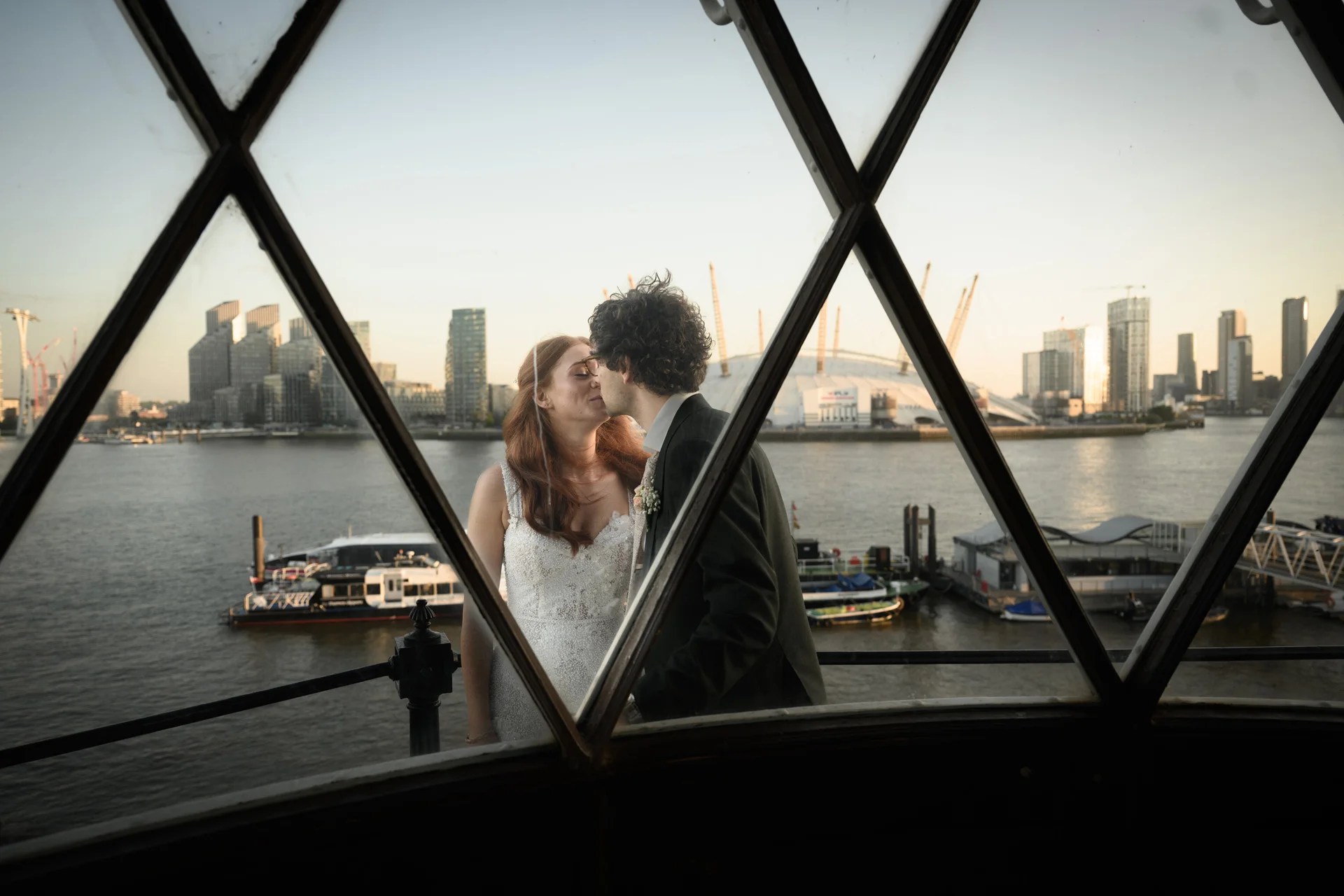 A newlywed couple share a tender kiss framed through the geometric window panes of a riverside venue overlooking the London skyline.