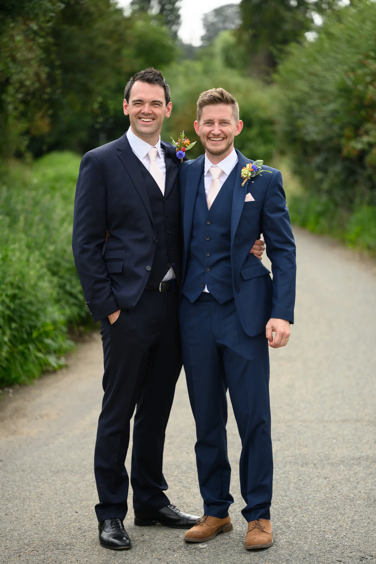 Two smiling grooms in navy three-piece suits with floral boutonnieres stand side by side on an outdoor path.
