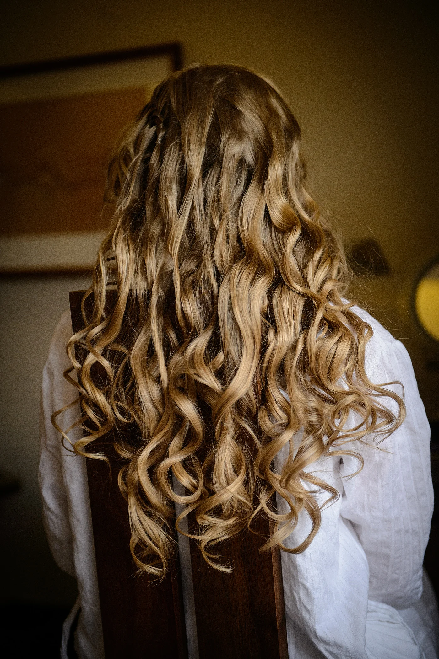 A close-up back view of a bride getting ready, showcasing her long blonde hair styled into soft, flowing curls while wearing a white robe.