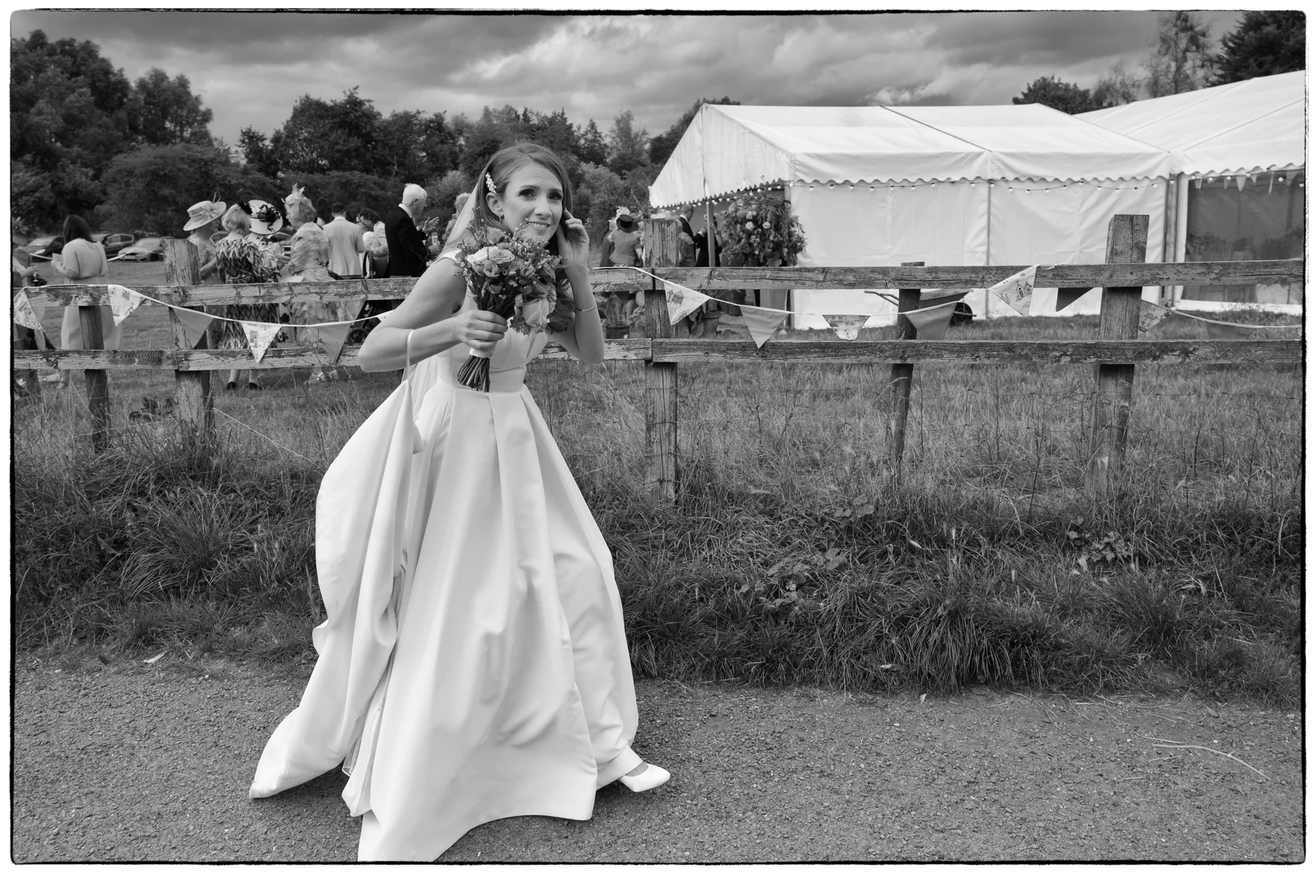 A joyful bride in a satin a-line dress smiles while holding a bouquet outdoors near a marquee decorated with bunting.