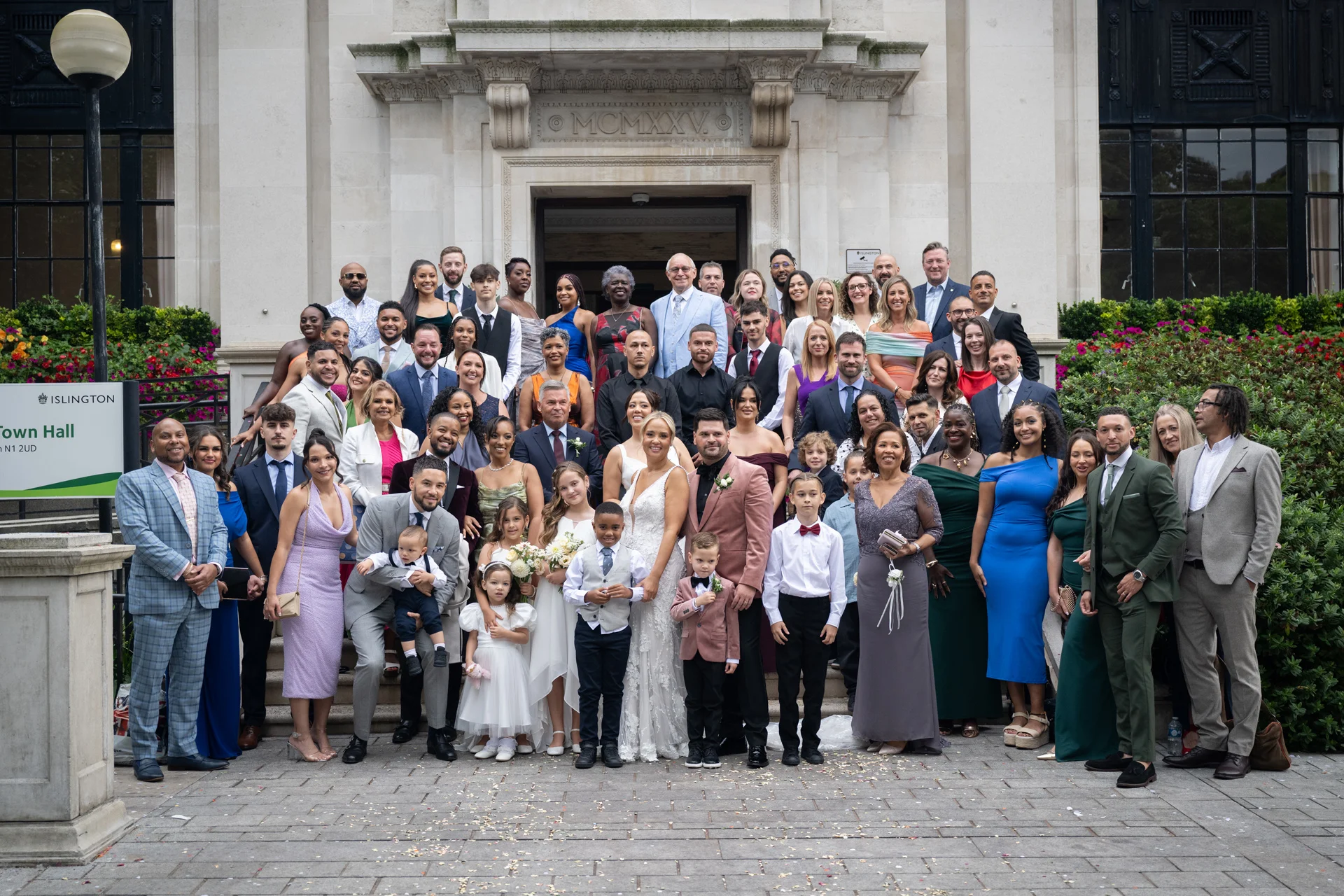 A large wedding party and guests pose for a group photograph outside the stone entrance of Islington Town Hall on a sunny day.