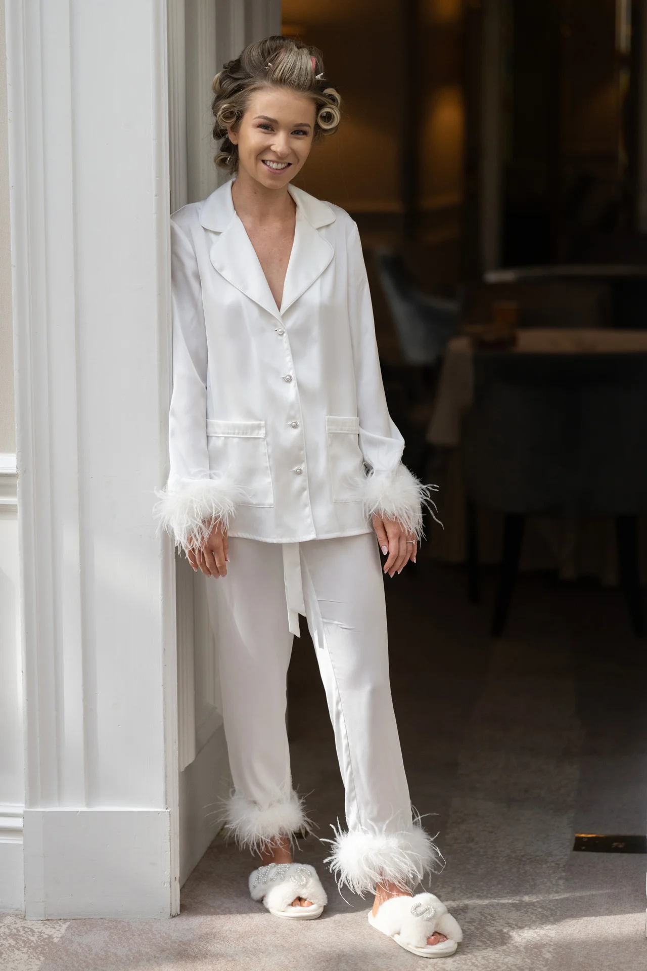A smiling bride wearing white satin pyjamas with feather cuffs and hair rollers leans against a white door frame during morning preparations.