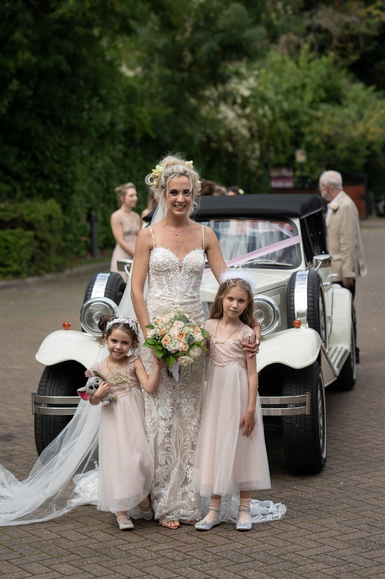 A smiling bride holding a floral bouquet poses in front of a vintage wedding car alongside two flower girls in matching pink dresses.