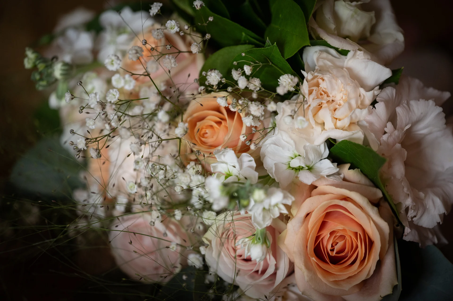 A detailed close-up shot of a hand-tied bridal bouquet featuring peach roses, pale pink carnations, and delicate baby's breath.