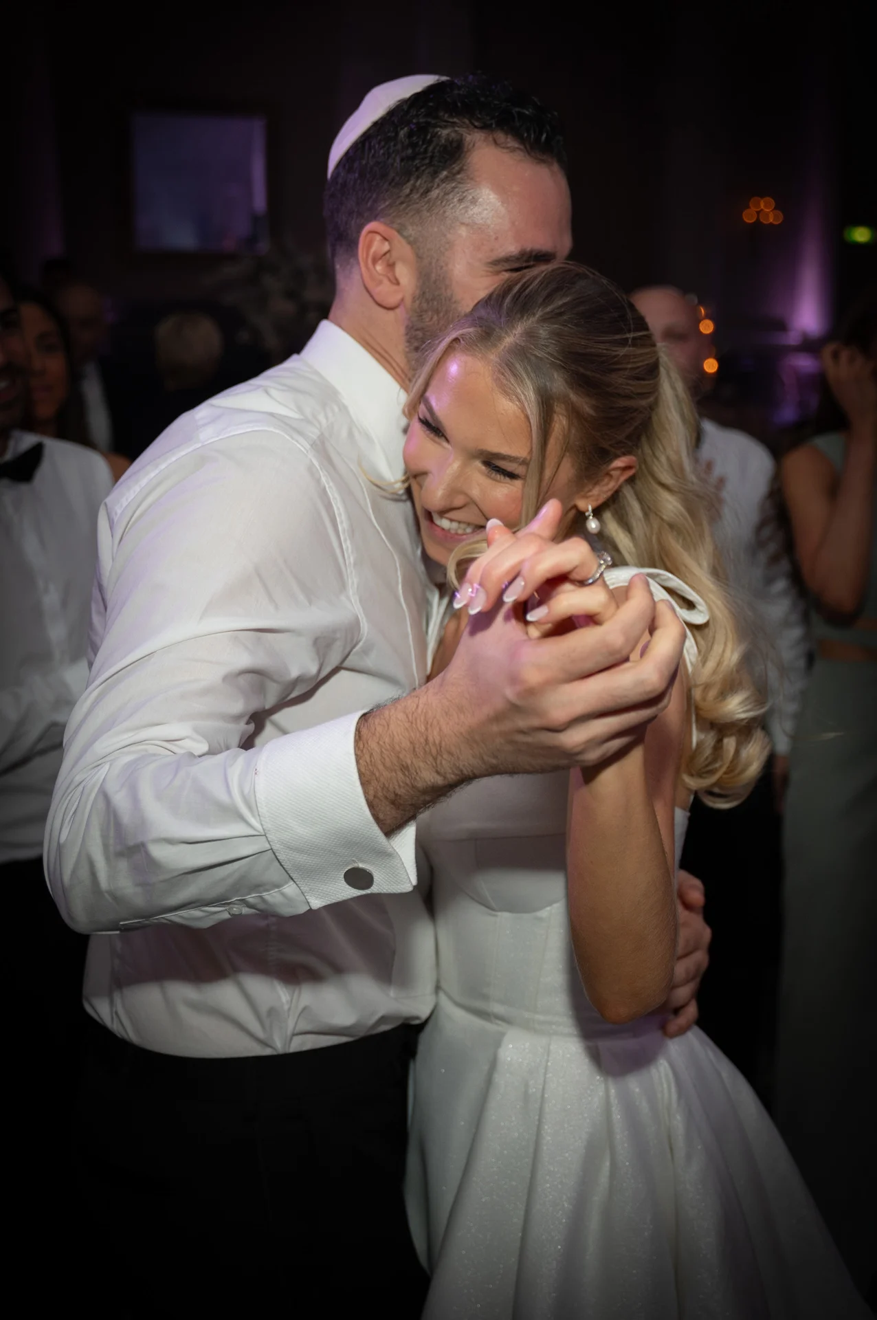 A newlywed couple shares a joyful first dance together on the dance floor, holding hands and embracing with bright smiles.